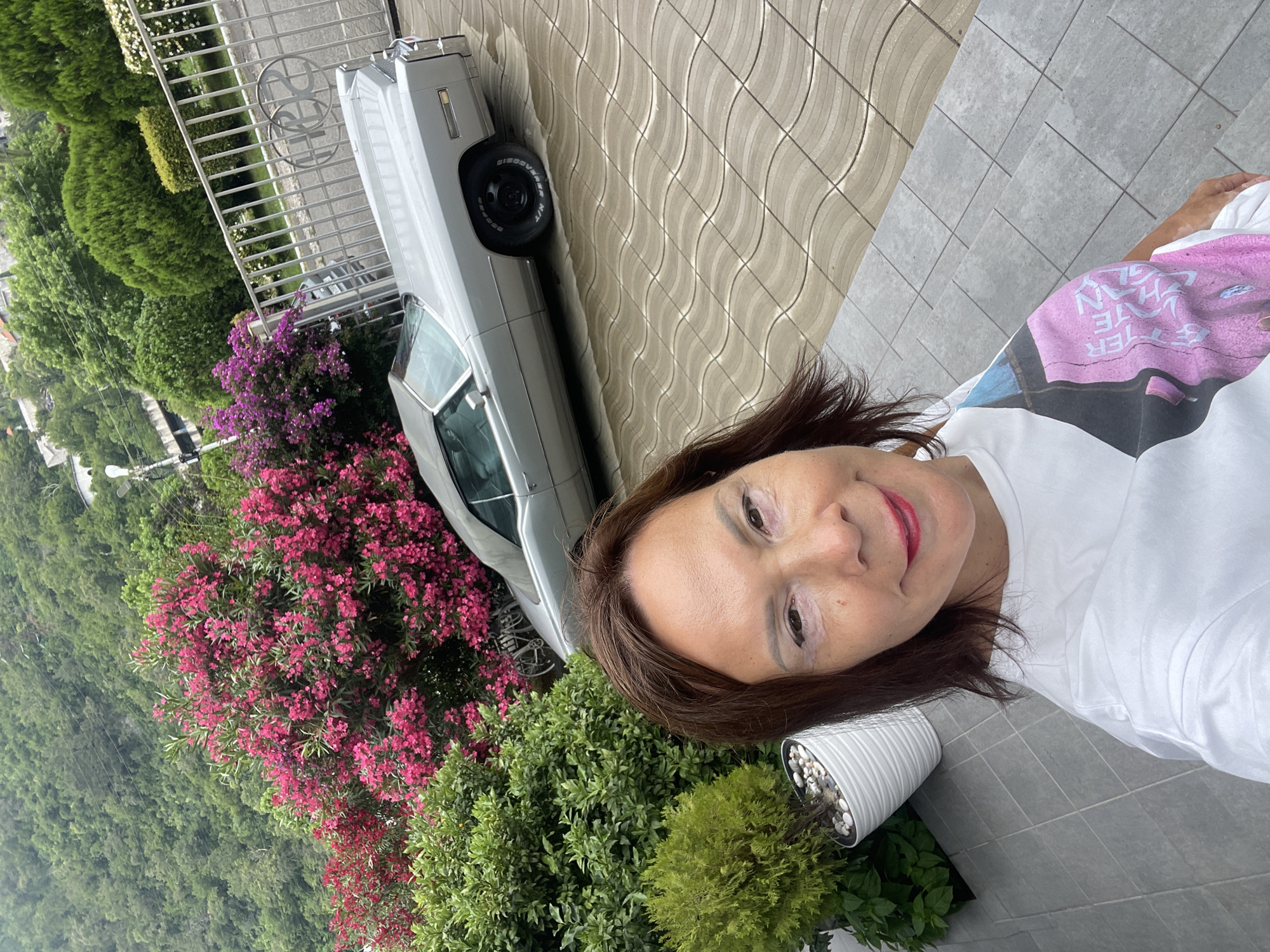 A woman with brown hair and red lipstick is taking a selfie outdoors. She is wearing a white t-shirt with a colorful graphic on it. To her left, a silver classic car is parked on a paved driveway. Behind the car, a lush green hillside covered with trees is visible. In the foreground, there are vibrant pink flowering bushes and manicured green shrubs. A white planter with small stones and some greenery sits to the woman's right. The scene appears to be set in a residential area with a fence and decorative metalwork in the background. The lighting suggests it is daytime, possibly a bright but overcast day due to the soft shadows.