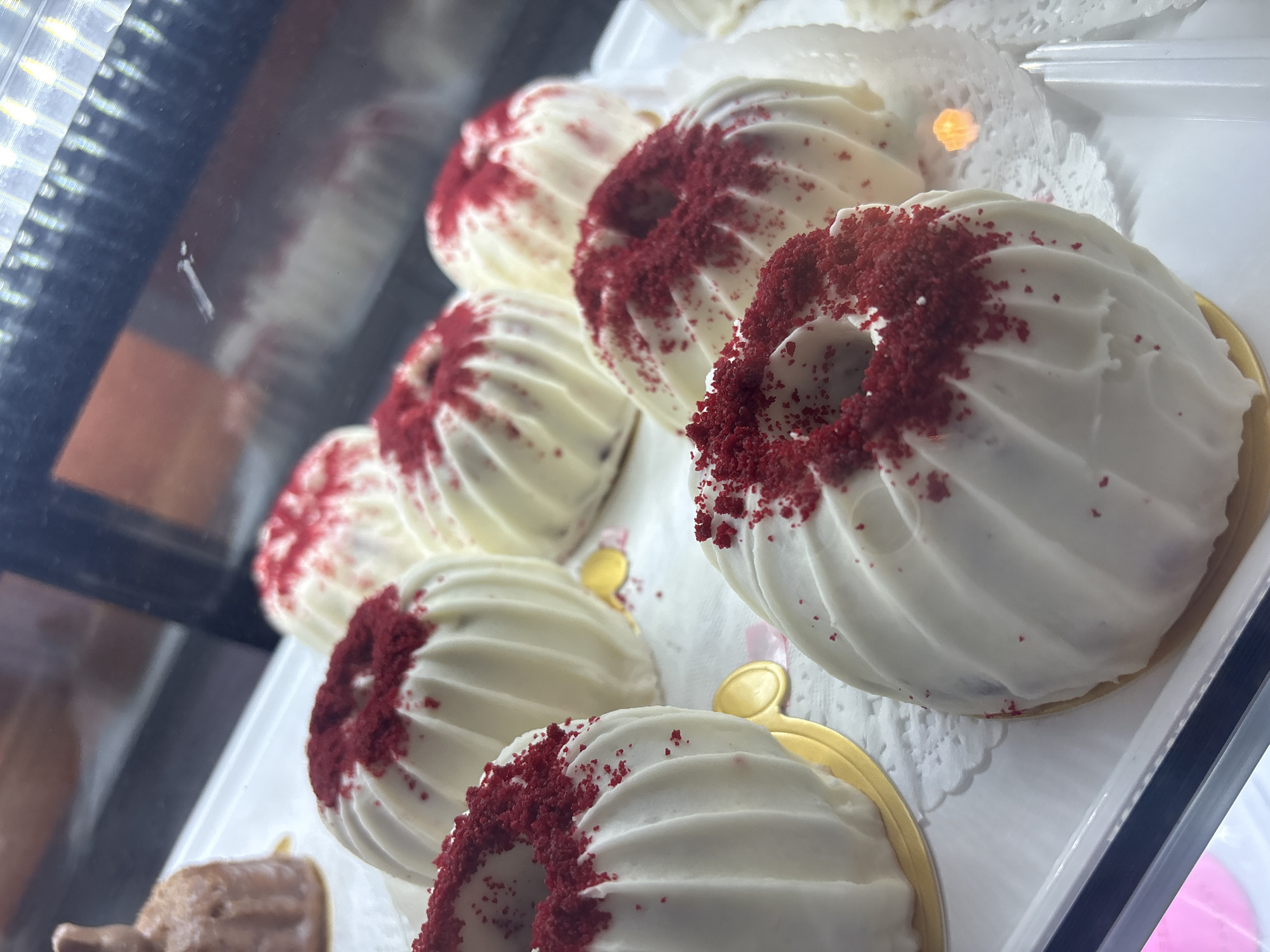 The image shows a display of red velvet bundt cakes in what appears to be a bakery or dessert shop. The cakes are arranged in rows on a tray, likely inside a refrigerated display case. Each cake is white with a red velvet crumb topping in the center. The cakes are placed on small, doily paper liners and some on gold plates. The cakes are fluted or swirled, and the red crumbs are scattered around the ring shape in the center of the cakes. The photo appears to be taken from the outside of the display case. While Puebla, Mexico is mentioned, there is no context to suggest the image originates from there.