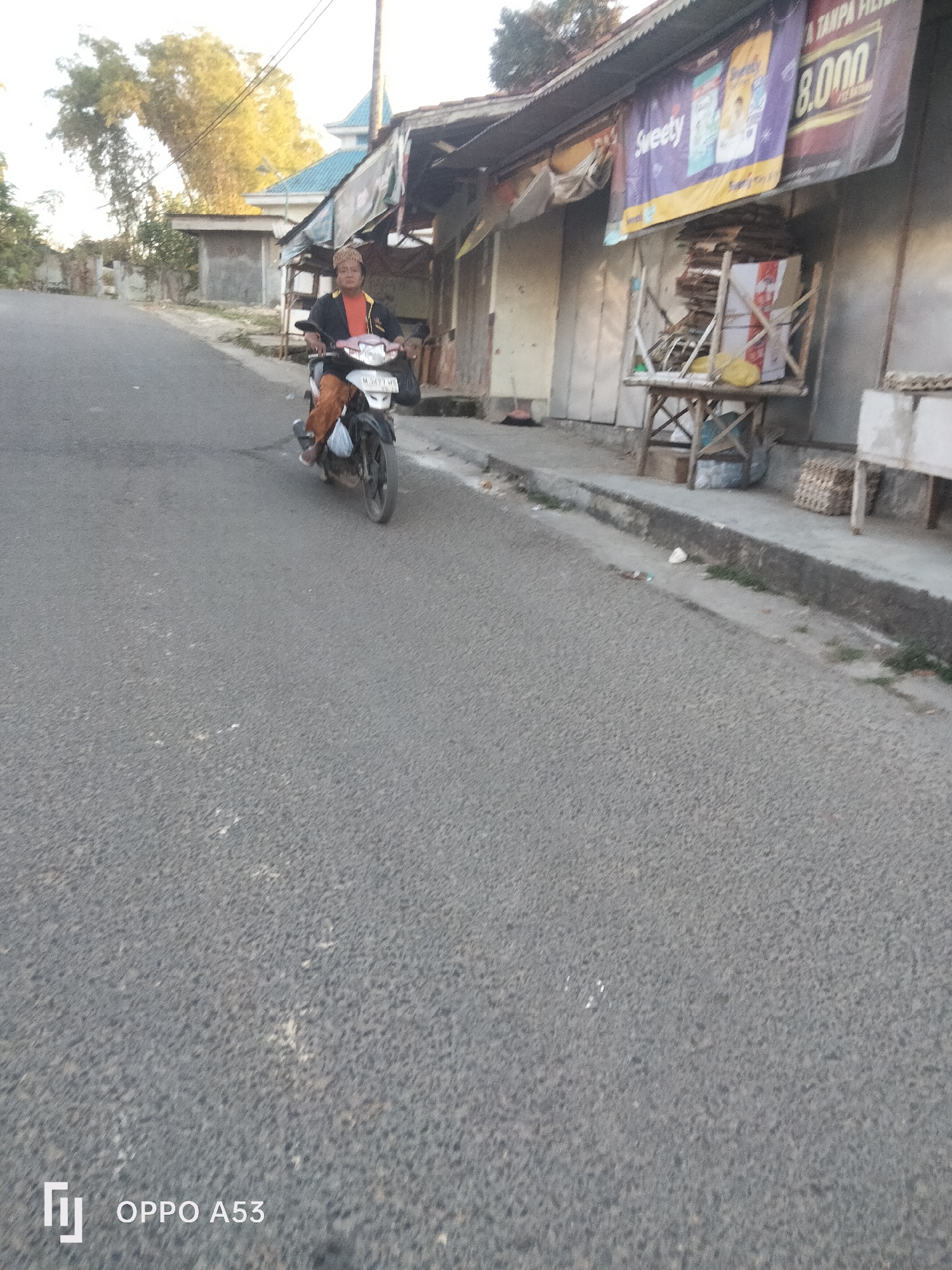 A person is riding a motorcycle on a paved road in Badung, Indonesia. The person is wearing an orange shirt and brown pants, and a patterned head covering. The motorcycle is black with a white front fender. The road is bordered by a sidewalk and buildings. One building has a sign that reads "Sweety" and "8.000". There are several boxes stacked on a wooden table in front of the building. The sky is clear and the sun is shining.