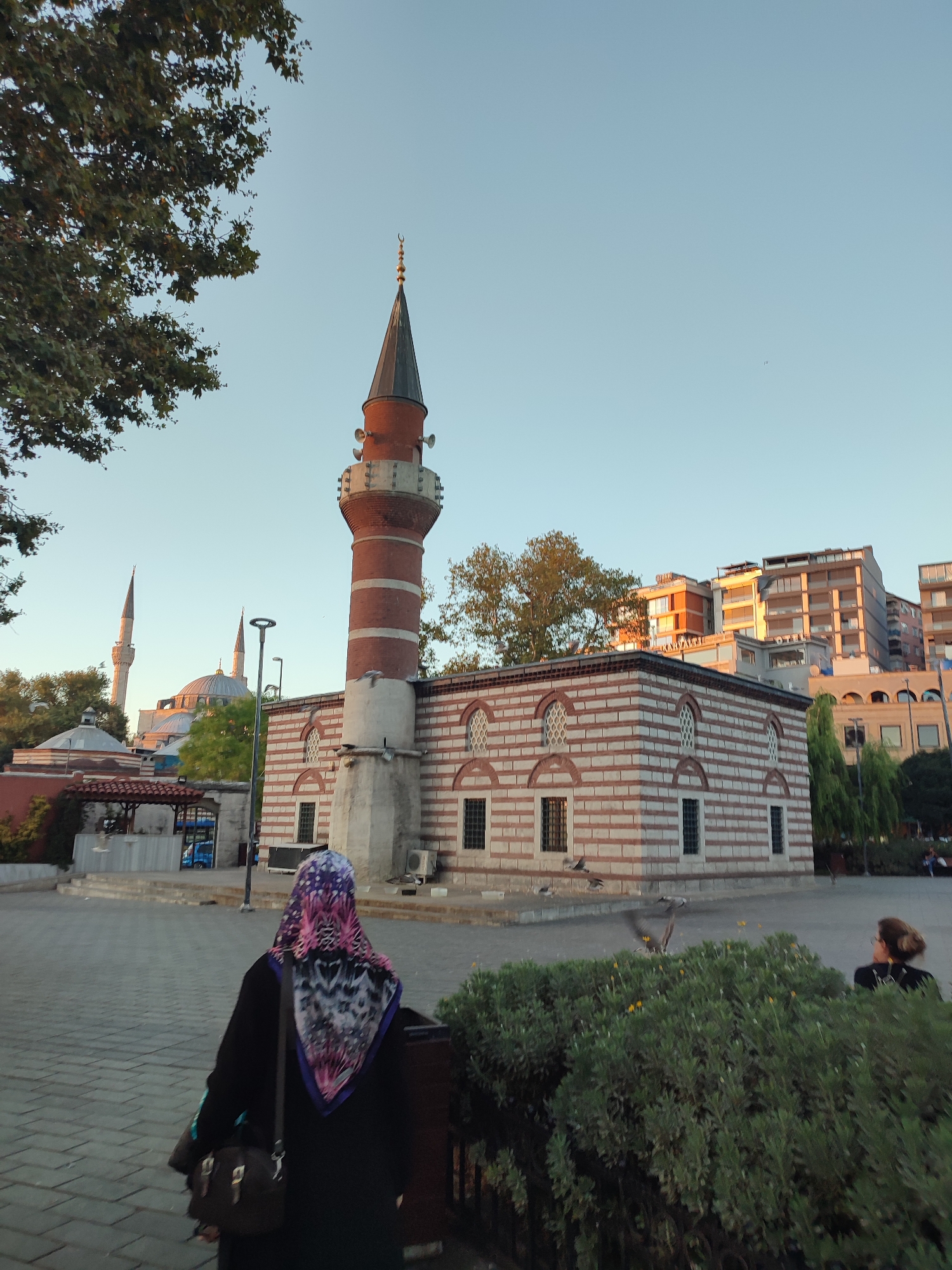 In Üsküdar, Turkey, a mosque with a red and white striped facade and a tall, slender minaret stands under a clear, pale blue sky. Sunlight illuminates the building, casting long shadows. In the foreground, a woman wearing a black abaya and a vibrant, patterned hijab walks away from the camera on a paved plaza. She carries a dark shoulder bag. Behind her, a lush green bush with small yellow flowers is visible. To the right, another woman, her back to the camera, sits on a bench, looking towards the mosque. Several pigeons are in flight across the plaza, some landing near the building. In the background, other buildings and minarets are visible, suggesting an urban setting.