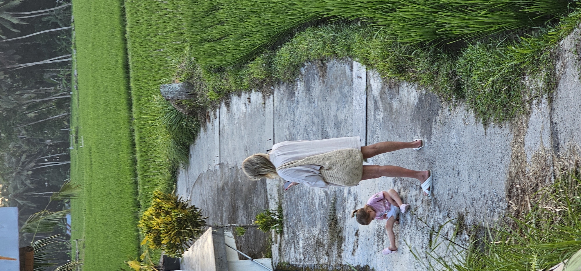 The image shows a woman and a young child standing on a concrete walkway bordered by lush green foliage in Tegallalang, Indonesia. The woman, fair-skinned with blonde hair, is wearing a light-colored dress and white sandals. She carries a woven bag on her shoulder and looks at her phone. The child, also fair-skinned with light hair styled in pigtails, wears a pink and white outfit with pink sandals, and is looking down towards the ground. The walkway is surrounded by vegetation, including rice paddies on one side and dense green grass on the other. In the background, there are tall trees with palm trees. The overall impression is one of a rural, tropical setting.