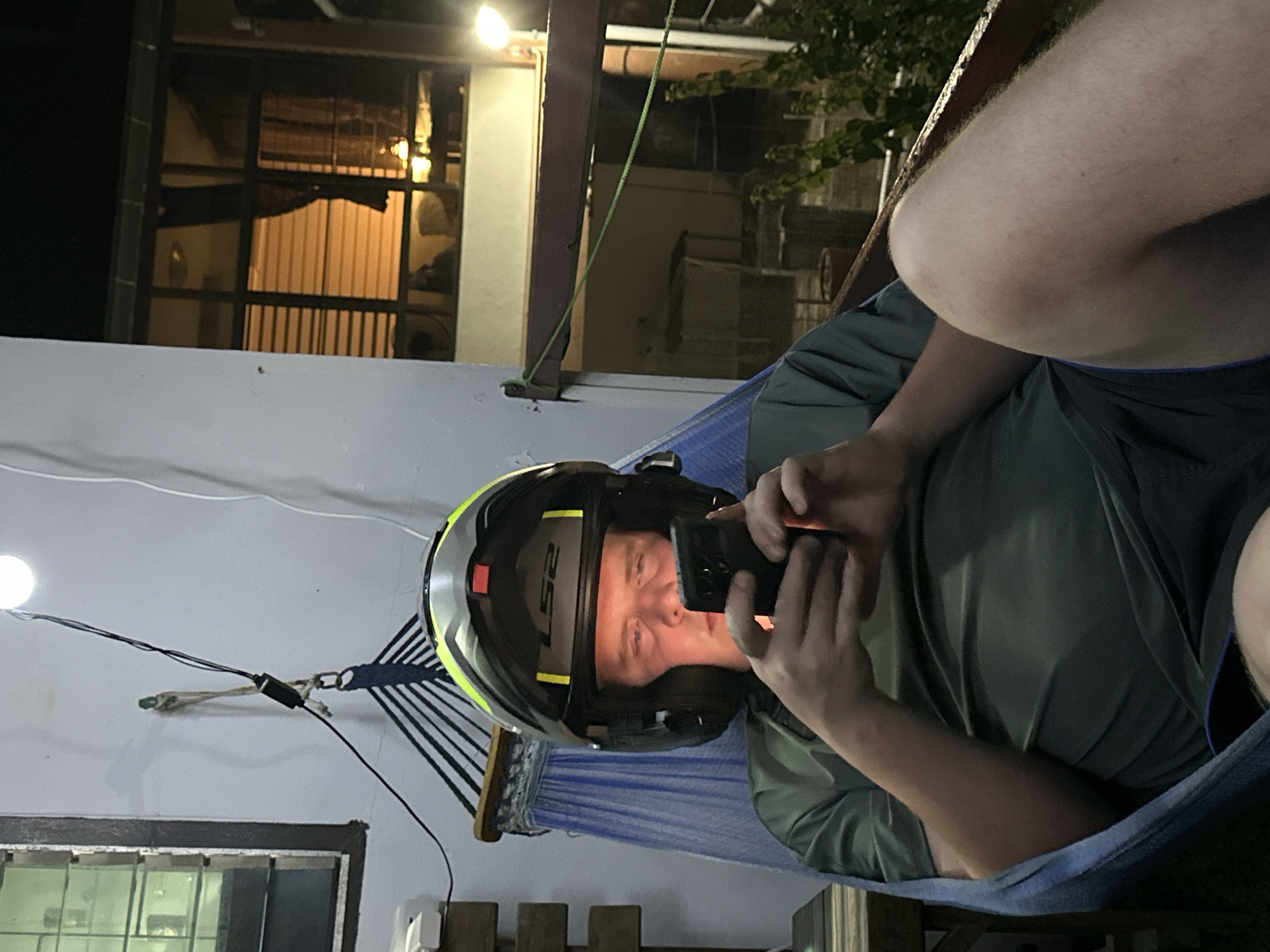 A light-skinned male wearing a green t-shirt, dark shorts, a black and yellow motorcycle helmet, and headphones is lying in a blue hammock. He is holding a black smartphone in both hands and looking at it. The scene appears to be outdoors at night, with a light bulb hanging from a wire above and to the left of the man. In the background, there is a window with blinds and interior lighting, suggesting a building or residence. The environment includes some vegetation and a railing. The overall setting suggests a casual, possibly residential, outdoor area in Karon, Thailand, during the evening or nighttime.