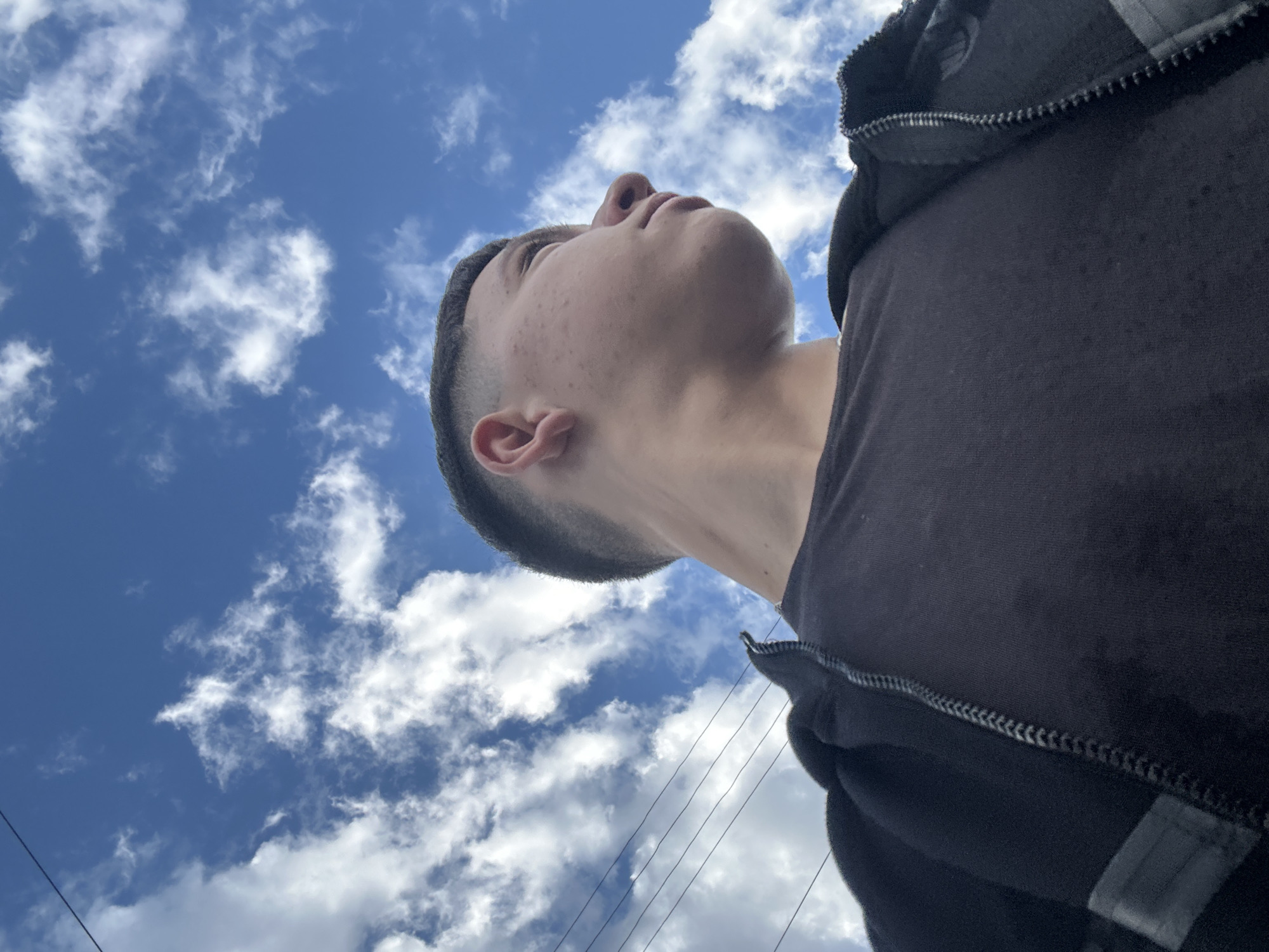 Low-angle shot of a young man with a short haircut, wearing a black jacket and t-shirt. He is looking upwards at a blue sky with scattered white clouds. Power lines are visible in the bottom of the frame. Location is undetermined in Zlatograd, Bulgaria.