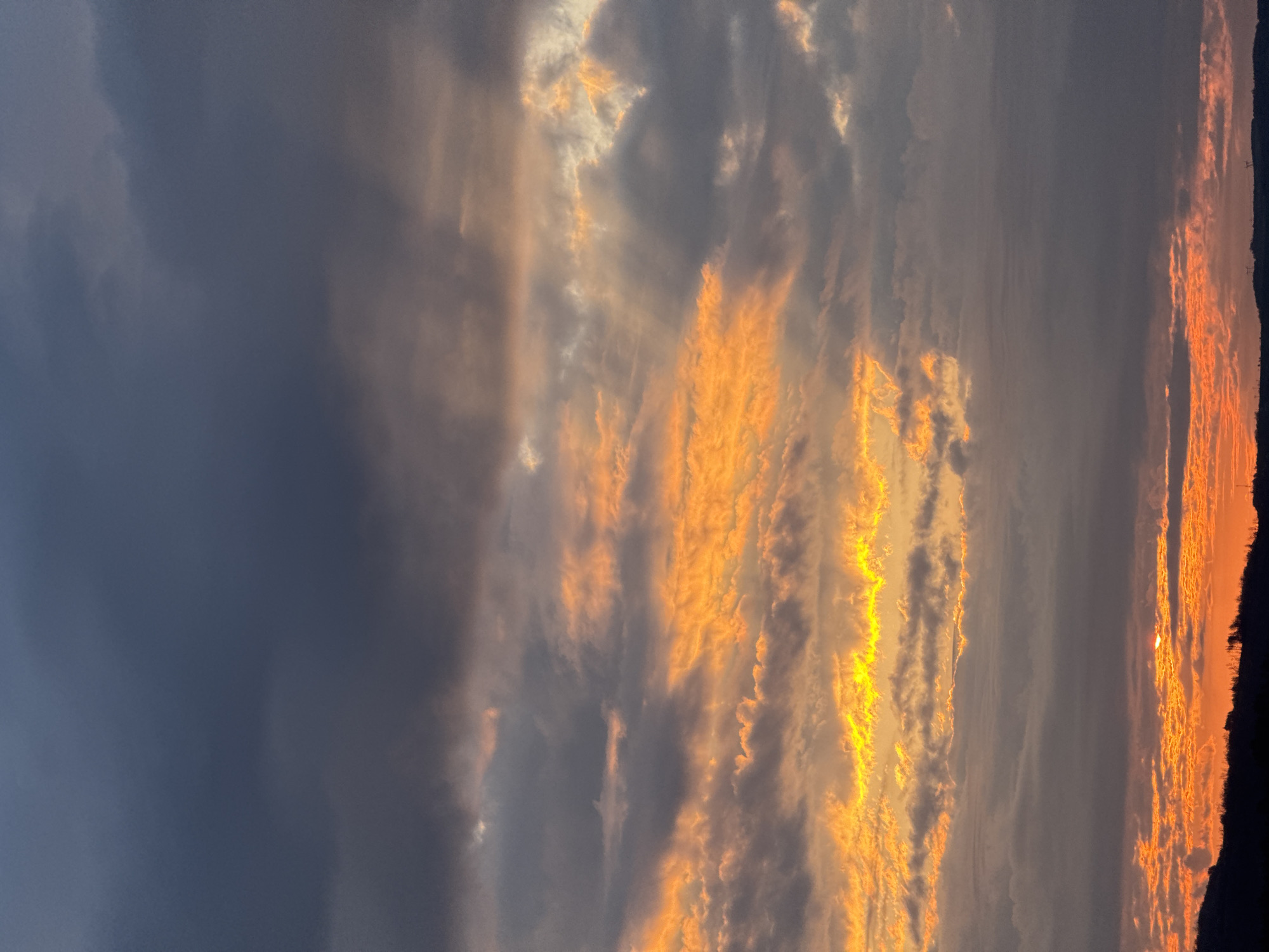 The image shows a cloudy sunset in Veliko Tarnovo, Bulgaria. To the left of the image are dark storm clouds. To the right of the image are thinner clouds with a golden glow. A dark strip of land is visible at the bottom right of the image.