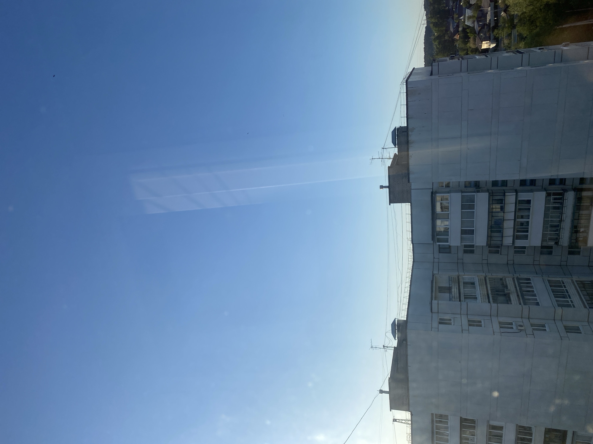 The image shows an upward view of a multi-story apartment building in Chaikovsky, Russia, juxtaposed against a clear blue sky. The building is constructed of concrete panels and features numerous windows and balconies. Several antennas are visible on the roof. The angle suggests the photo was taken from a nearby, potentially slightly elevated, position. The sky is mostly clear with some faint contrails or cloud formations. A small, dark bird is visible in the upper left quadrant of the sky. A blurred area in the sky might indicate window reflections. Beyond the apartment building, treetops and rooftops indicate a residential area. Power lines run alongside the building.
