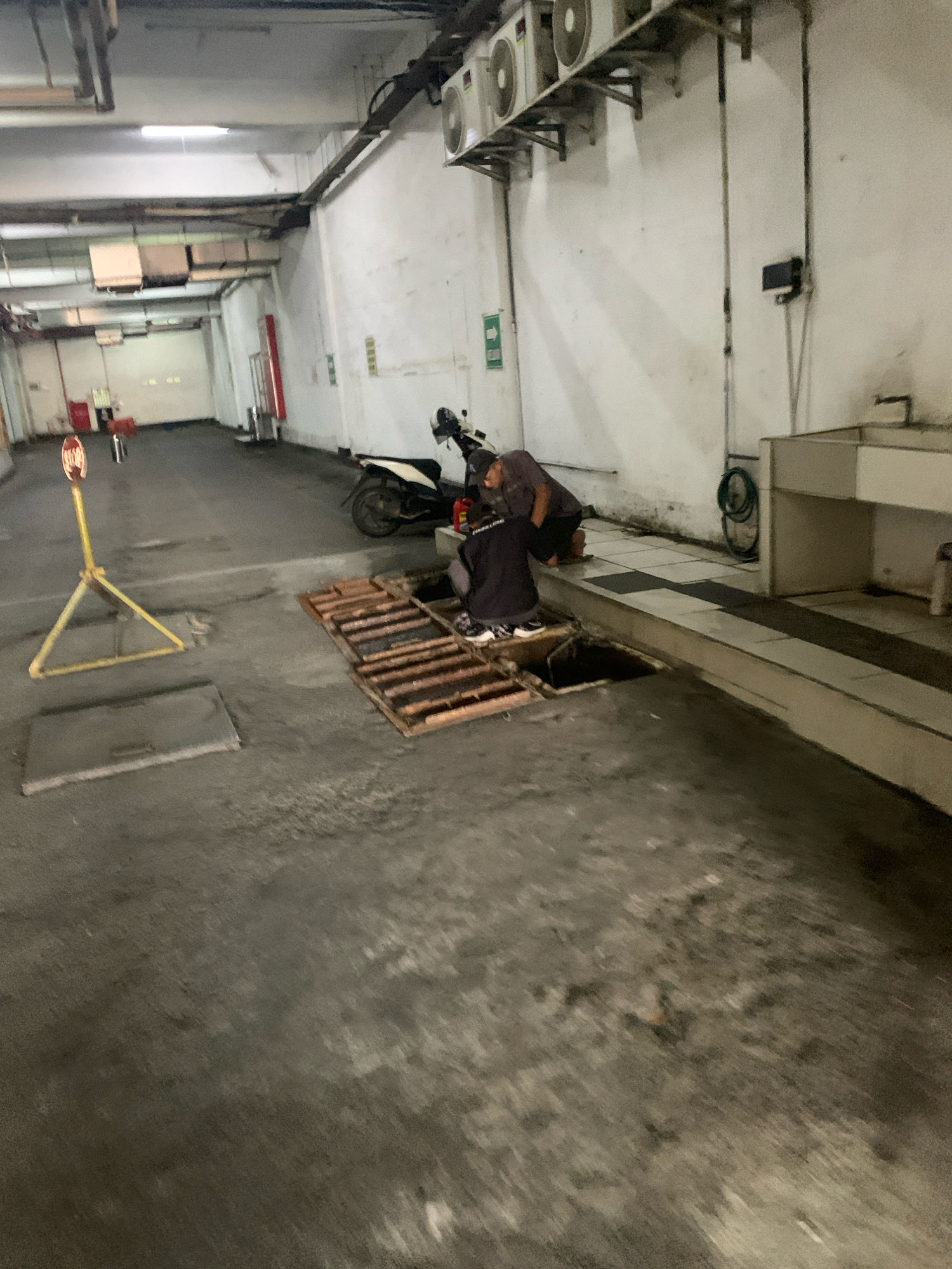 The image depicts an outdoor, possibly covered, area that appears to be a maintenance or workshop space in Malang, Indonesia. Two individuals are observed working near an open drainage grate. One person, wearing a dark shirt and pants, is crouched down looking into the grate. Another individual, wearing a grey shirt and dark pants, is also crouched nearby, possibly assisting or observing. A white and black motorcycle is parked behind them. In the background, the environment consists of concrete walls and flooring, with air conditioning units mounted high on the wall. A yellow cautionary sign on a stand is visible to the left, along with a closed manhole cover on the ground. The overall impression is of a functional, utilitarian space.