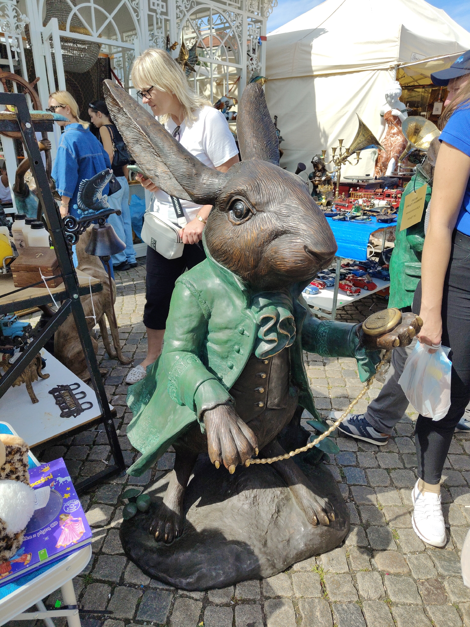 The image depicts an outdoor market scene in Gdańsk, Poland, likely on a cobblestone street. In the foreground, a large bronze statue of a rabbit dressed in a green coat and waistcoat is the central subject. The rabbit is holding a pocket watch in its right hand and has a pearl necklace draped around its neck. A portion of a white shopping bag and a person's legs and feet, wearing white sneakers, are visible to the right of the statue. In the background, several people are browsing various market stalls. One woman with blonde hair and glasses is examining items at a stall. Another stall features a white tent, and on display are numerous small toy cars, a gramophone, and other decorative objects. The overall environment suggests a lively market day with a variety of vendors and shoppers.