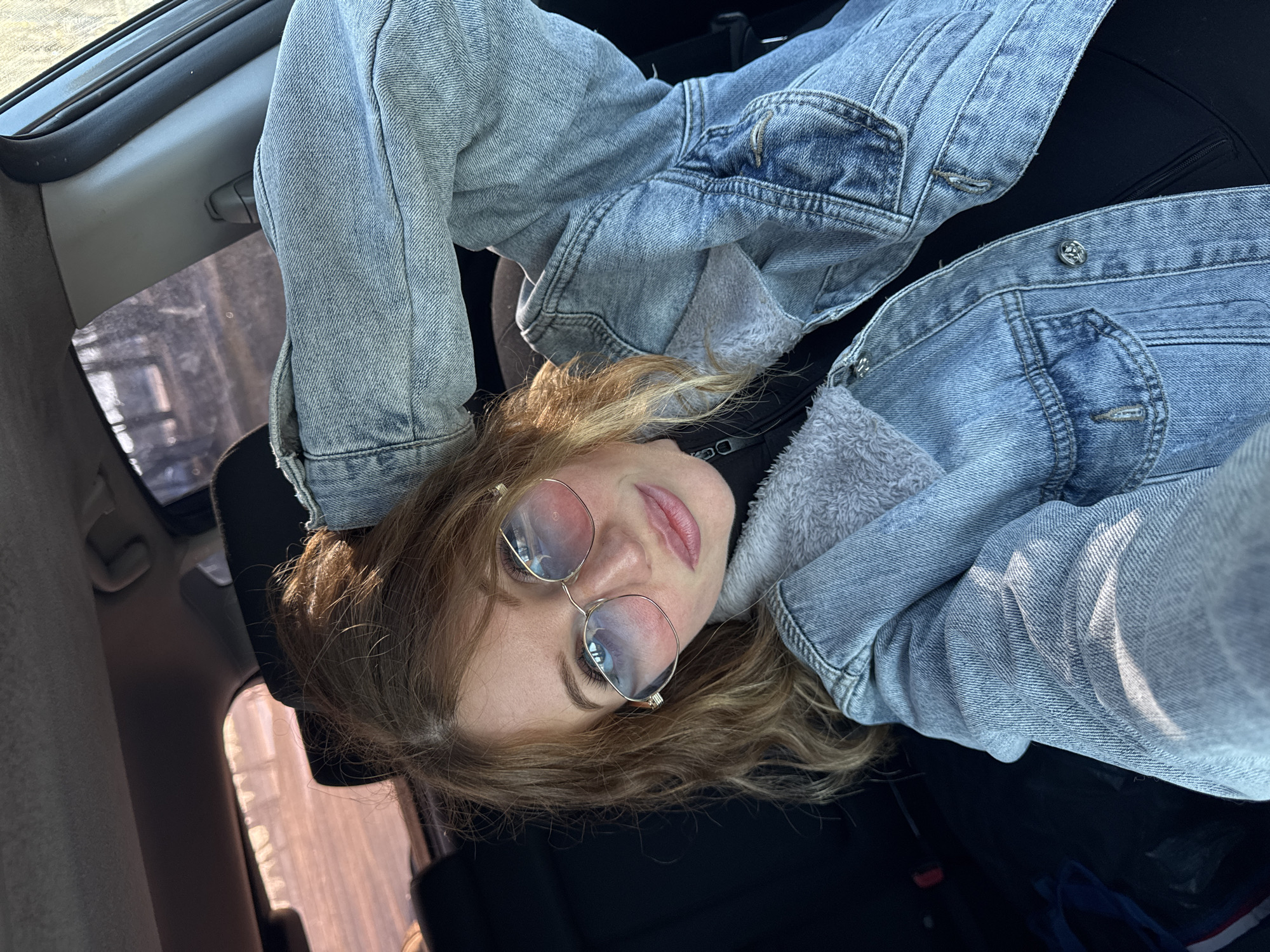 A young adult woman wearing a light blue denim jacket and round metal-rimmed glasses reclines in the passenger seat of a car. Her head rests against the headrest. The car's interior is black and gray. Through the window, there are buildings and possibly trees, suggestive of a residential or urban area, though the specific location in Noviy, Russia, cannot be determined from the image. There is no observable action or interaction.