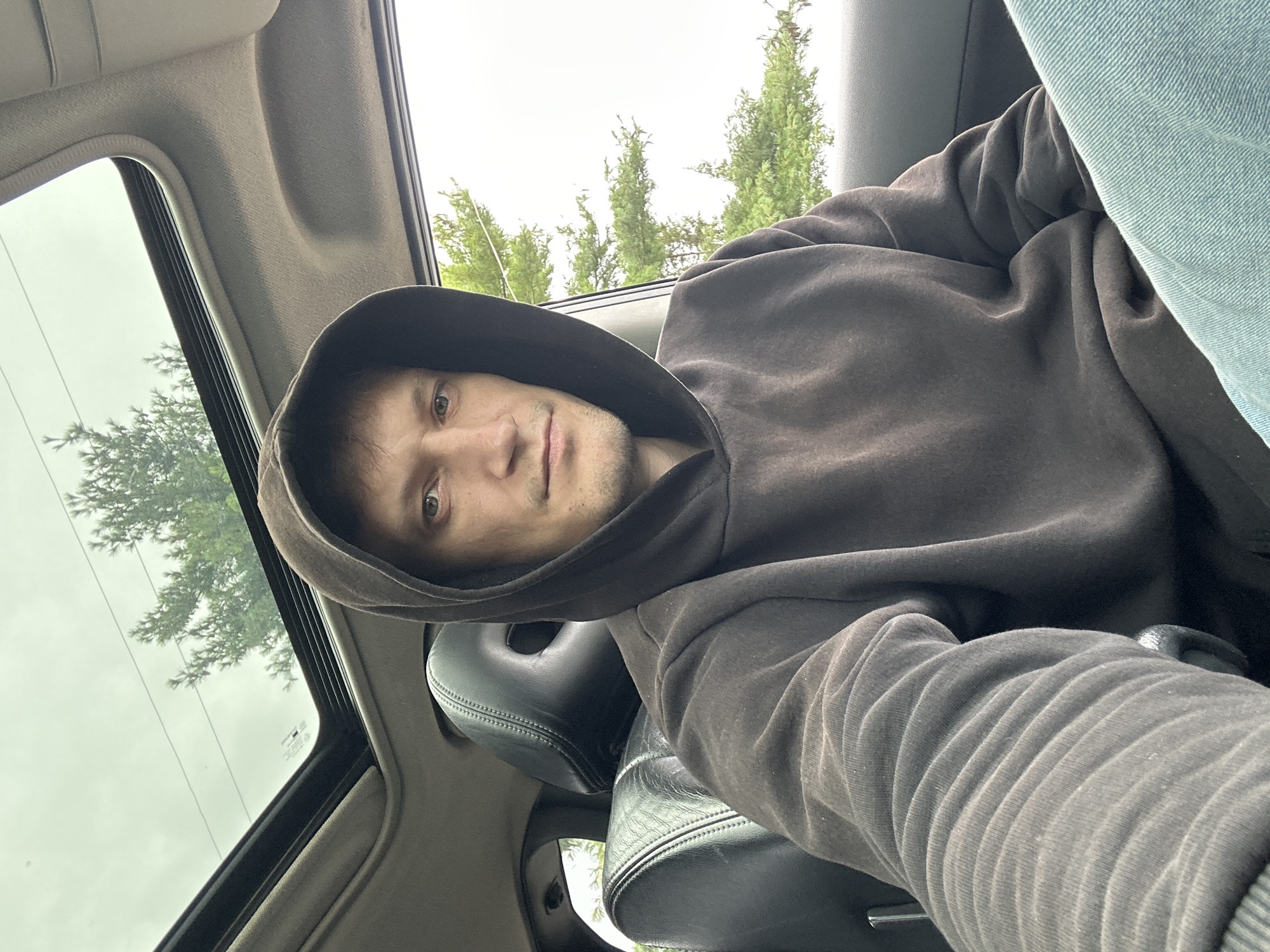 A man wearing a dark brown hooded sweatshirt and light blue jeans is sitting in the driver's seat of a car. The car has a sunroof which is open, revealing a cloudy sky and green trees above. The interior of the car is visible, including black leather seats and the dashboard. The man is looking at the camera with a neutral expression. The image appears to be taken during the day in an outdoor setting, possibly in the city of Makiv, Ukraine.