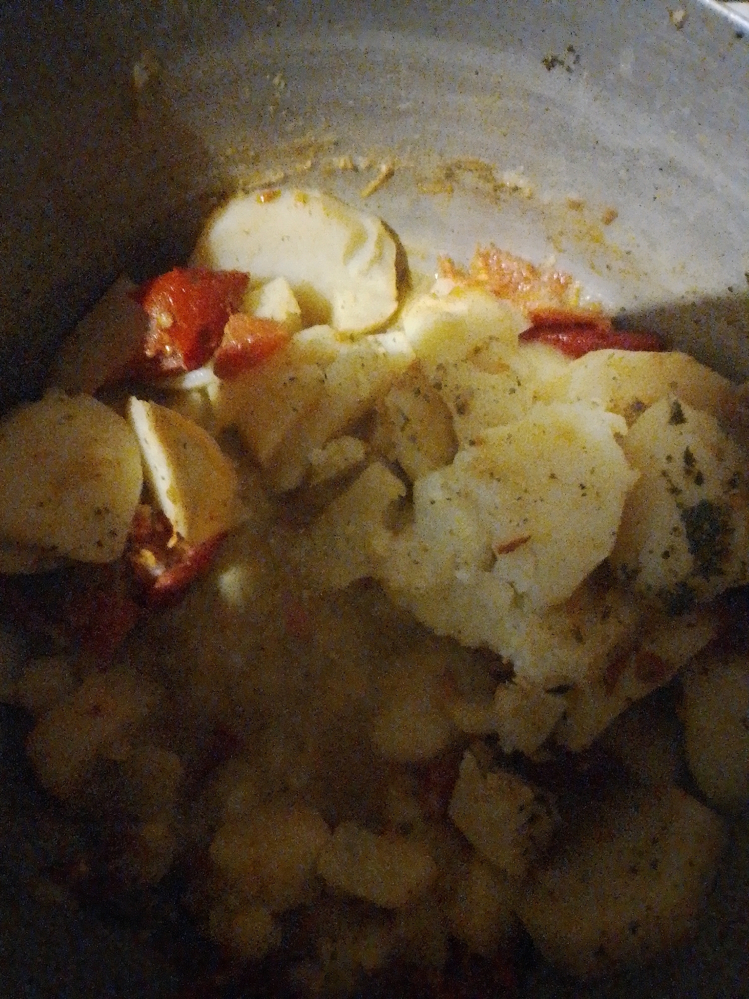 A close-up, overhead shot shows a pot of food, likely a stew, filled with potatoes and pieces of tomato. The potatoes are cut into irregular chunks and appear soft, some slightly browned. Red pieces of tomato are interspersed throughout, some appearing cooked down and blended with the sauce. The overall color palette is warm, with dominant yellows, browns, and reds. The pot itself is a dull metallic color, showing signs of use. The lighting is dim, creating shadows and highlighting the texture of the food. The scene appears to be an indoor kitchen setting, possibly in Myrhorod, Ukraine, given the provided context. There are no people visible in the image.