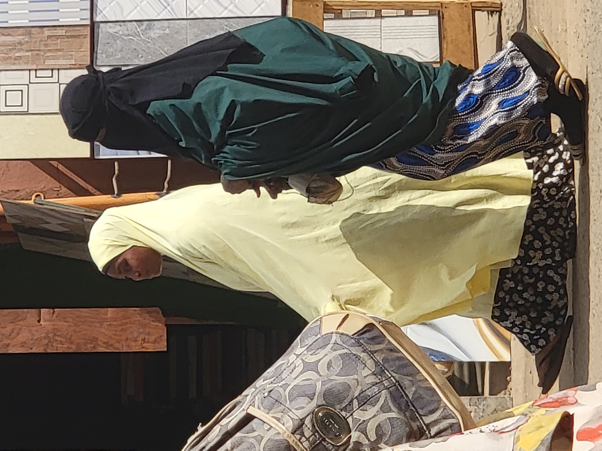 Two women are standing next to each other. One is wearing a black head covering, a green shawl, and a blue and white patterned skirt. The other is wearing a light yellow head covering and a black and white patterned skirt. A patterned handbag is visible in the foreground. The background includes various tiles displayed on a wall. The location is identified as Maiburiji, Nigeria.