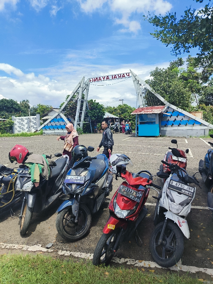 The scene is a parking lot located in Majalengka, Indonesia at the coordinates -6.846, 108.204. The lot is paved with gray asphalt. There are six motorcycles parked on the lot. Two men and one woman are standing in the parking lot. The woman is wearing a pink headscarf and a white blouse. The men are wearing jeans and short-sleeved shirts. One man is wearing a blue jacket. Behind the motorcycles, a metal archway with the words "MAYA IAJAH" written on it in reversed letters is visible. The archway is situated in front of a small blue structure. In the distance, behind the motorcycles, a group of people can be seen walking on the road. The sky is bright blue and partly cloudy. There are green trees in the background, and the scene is well lit by the sun.