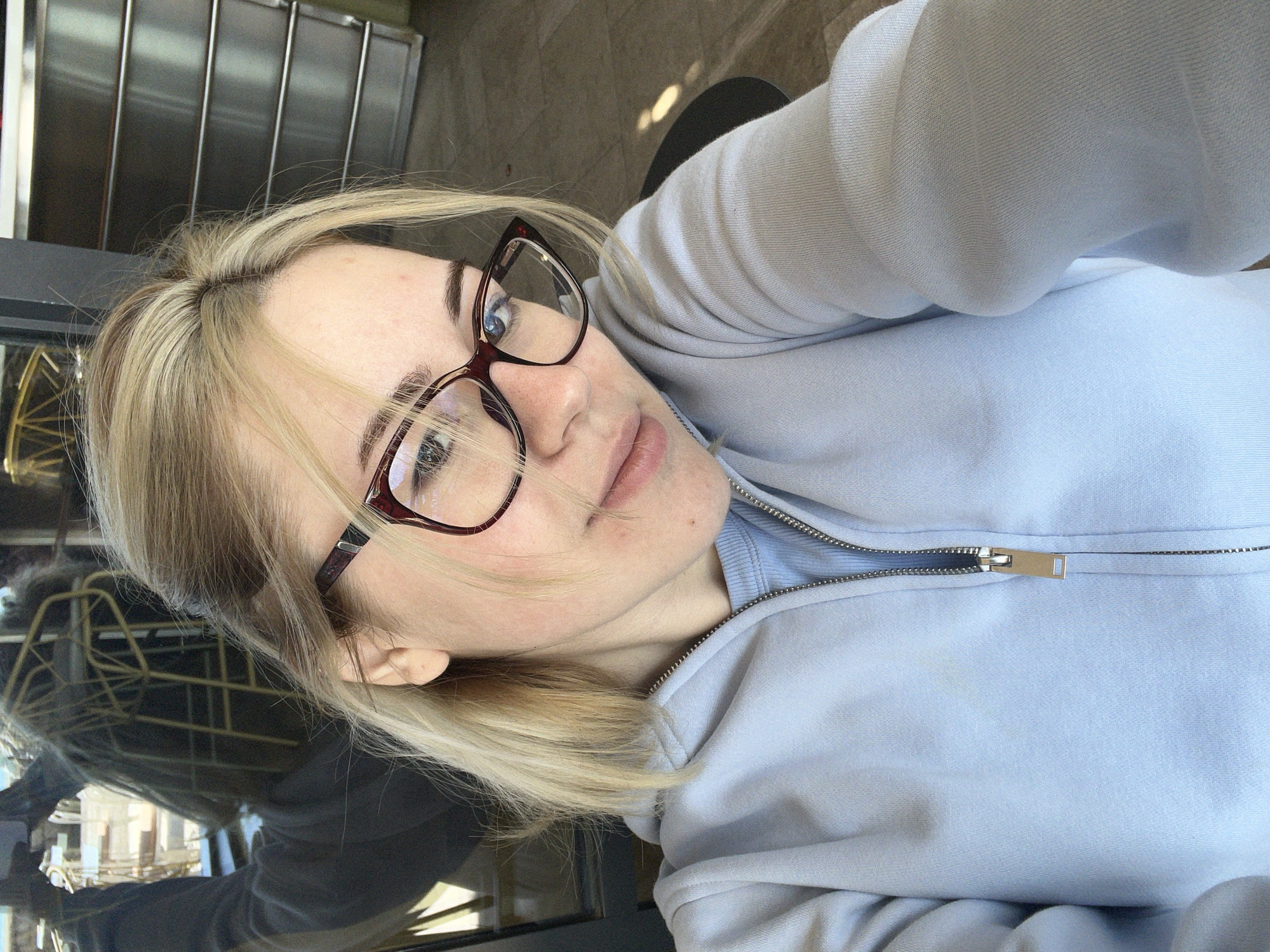A light-skinned woman with blonde hair and glasses is posing in a selfie. She is wearing a light blue zippered sweatshirt. The background reflects a building or structure with metal bars and other objects. The location is in Rafailovići, Montenegro, evidenced by a blurry reflection of a building with scaffolding.