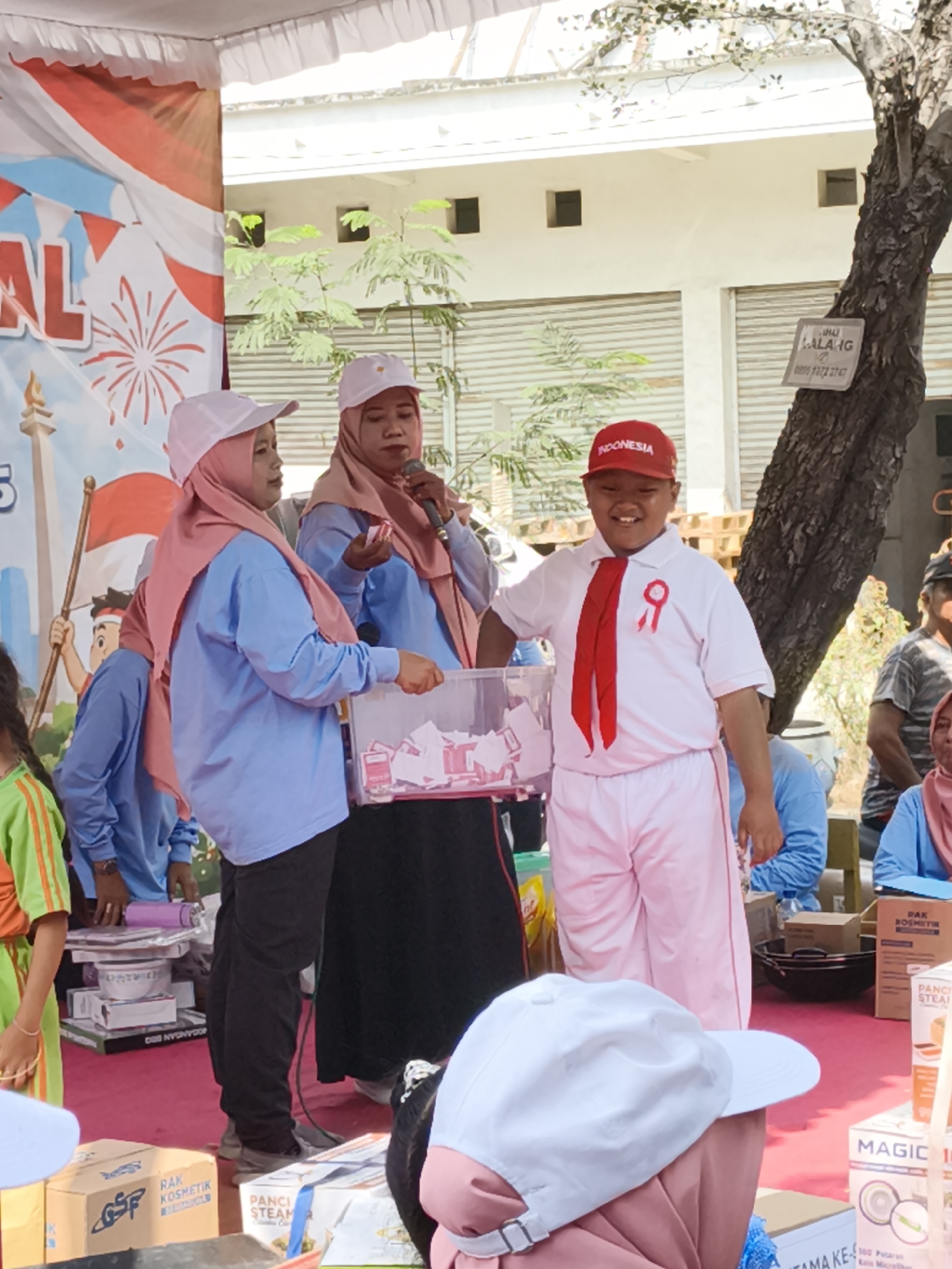 In a brightly lit outdoor event space in Petiken, Indonesia, a group of people are gathered around a stage. A young boy, wearing a white uniform with a red tie and a red cap that reads "INDONESIA," beams with a wide smile. He is positioned in front of a clear plastic bin filled with folded papers, suggesting a lottery or raffle. To his left, two women in pink hijabs and light blue shirts stand close. One woman holds a microphone and appears to be speaking, while the other is reaching into the bin. Behind them, a colorful banner with red and white stripes and fireworks imagery is visible, along with part of a building with a corrugated metal door and small square windows. The ground is covered with a bright red carpet, and various boxed items, including ones labeled "RAK KOSMETIK" and "Panci Steam," are arranged on tables and the floor. The overall atmosphere seems festive and communal, possibly related to a local celebration or market event.