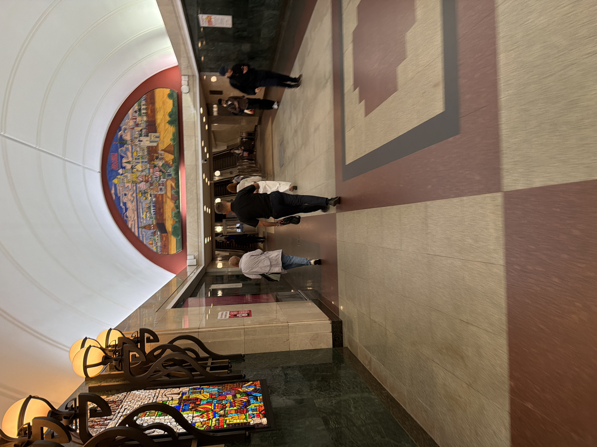 The image depicts an interior view of a Moscow Metro station, specifically the corridor leading to or from escalators. The architecture features a high, arched ceiling, a large, colorful mural depicting a cityscape with golden domes, and decorative lamps. The walls are adorned with green marble and a stained-glass panel. The floor is tiled with a pattern of beige and reddish-brown rectangles, with darker inlaid borders. Several individuals are walking through the station. In the foreground, a man in a black shirt and dark pants walks away from the camera, carrying a black bag. Further back, a woman in a light-colored jacket and jeans walks in the same direction. Other individuals are visible in the background near the escalators, including a man in a dark jacket and cap looking at his phone. The overall atmosphere is that of a busy public transportation hub.