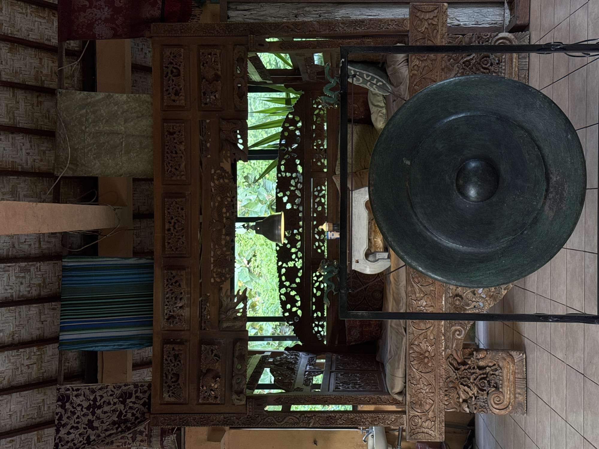 The image shows an interior scene in Kuta Selatan, Indonesia, featuring a large gong centered in the frame, mounted on a black metal stand. The gong's surface has a visible center boss. Behind the gong is an intricately carved wooden structure that appears to frame a window or opening. Through this frame, lush greenery can be seen, suggesting a garden or natural environment outside. Various fabrics are draped around the room, adding a touch of color and texture. The setting is a tiled floor area. The image is photographed from a slightly low angle.
