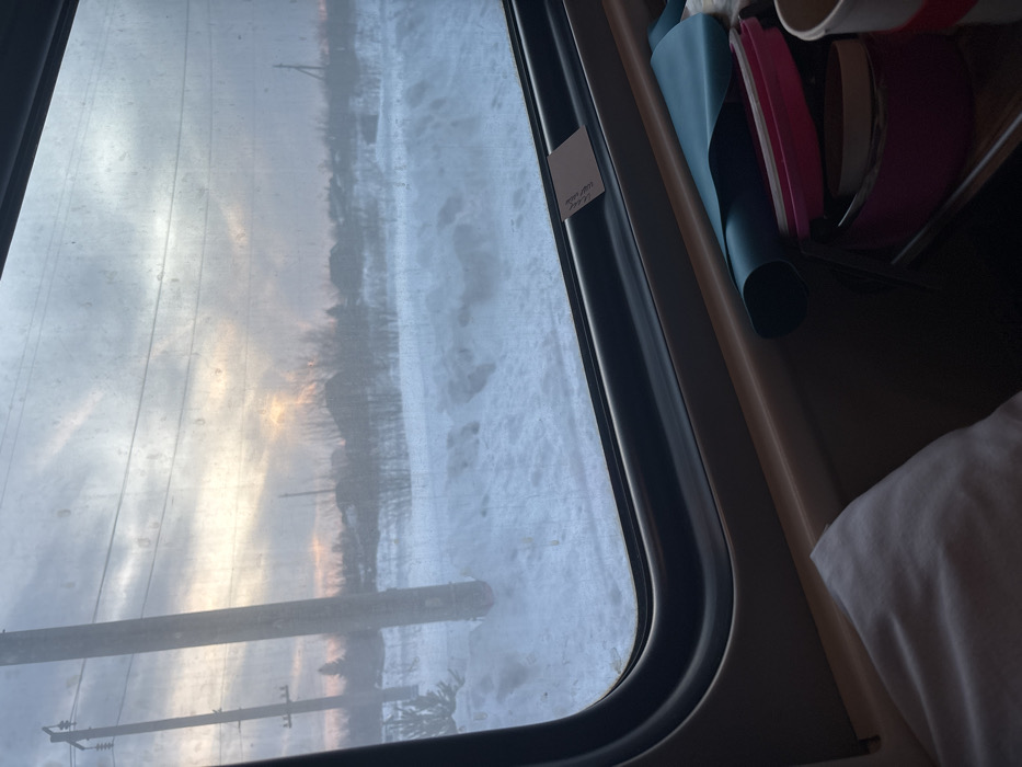 This image captures a winter landscape viewed from inside a moving train in Pervomaiskoe, Russia. The scene outside the dark-framed train window displays extensive snow cover across uneven terrain. Utility poles with electrical wires traverse the midground, accompanied by sparse, bare trees and the silhouettes of distant buildings against the horizon. The sky above is overcast with grey clouds, from which a bright, localized yellow-orange light, indicative of a sunrise or sunset, emerges towards the upper-left, illuminating the cloud layers. Inside the train, a light brown window sill is visible, holding a small, light-colored card tucked into the frame. Below the sill, on an interior shelf, a teal rolled item is situated next to several stacked pink circular containers and a partially visible white disposable cup with a red band. A white fabric object, possibly a pillow or blanket, is partially visible in the bottom-left corner of the frame.