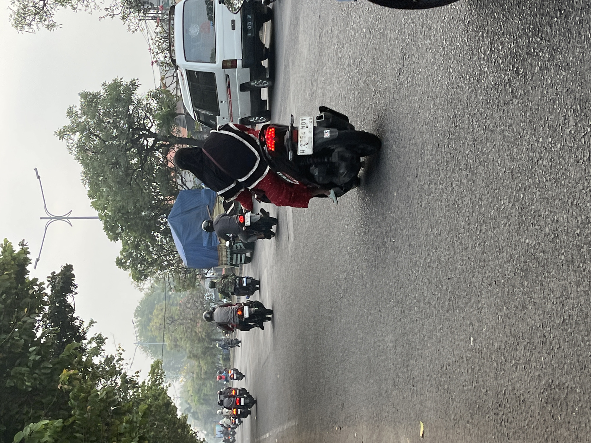The image depicts a street scene in Sumurame, Indonesia, likely during daylight hours with overcast skies. A procession of motorcycles is traveling down a paved road lined with trees. In the foreground, a person wearing a red dress and a black hijab is riding a black motorcycle, with its license plate visible as "W 2754 NDY". Behind this motorcycle, several other motorcycles are in motion, some with passengers. Further down the road, a white van with a red stripe and a blue tarp-covered truck are also visible. The environment suggests an urban or suburban setting, with the presence of vehicles and trees indicating a developed area.
