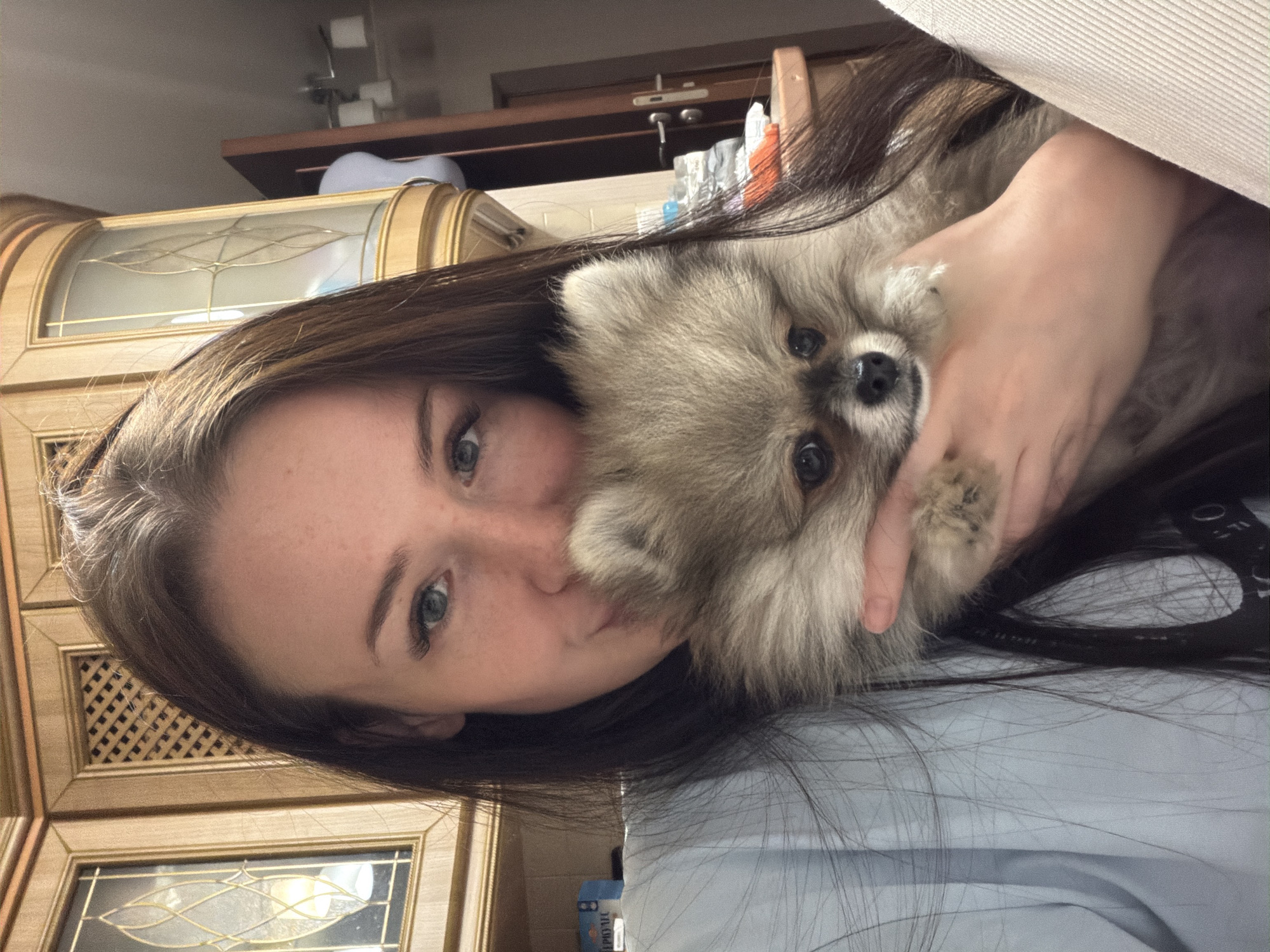 An image, rotated 90 degrees clockwise, captures a young woman holding a small, fluffy dog indoors, in what appears to be a home setting in Russia. The woman, with long dark hair cascading around her, light blue eyes, and fair skin with visible freckles, looks towards the camera with a relaxed or slightly pouting expression. She is wearing a light-colored garment, possibly a light blue shirt.
Cradled in her arms and close to her face is a Pomeranian-type dog with light brown and grey fur, dark eyes, and a small, black nose. The dog also looks directly at the camera, its expression calm. One of the woman's hands is gently supporting the dog's head.
The background features rich wooden cabinetry with decorative glass panels and gold-colored accents, suggesting a traditional interior design. Above the cabinetry, a wooden shelf holds a few indistinct items, and a white, possibly electronic device is visible. Part of a light-colored couch or upholstered furniture is visible in the upper right corner, against which the woman seems to be resting. Near the woman's left shoulder, in the background, a blue package or box is visible with the text "Murat" printed on it. The lighting is soft, suggesting an indoor environment, possibly with artificial light or indirect daylight.