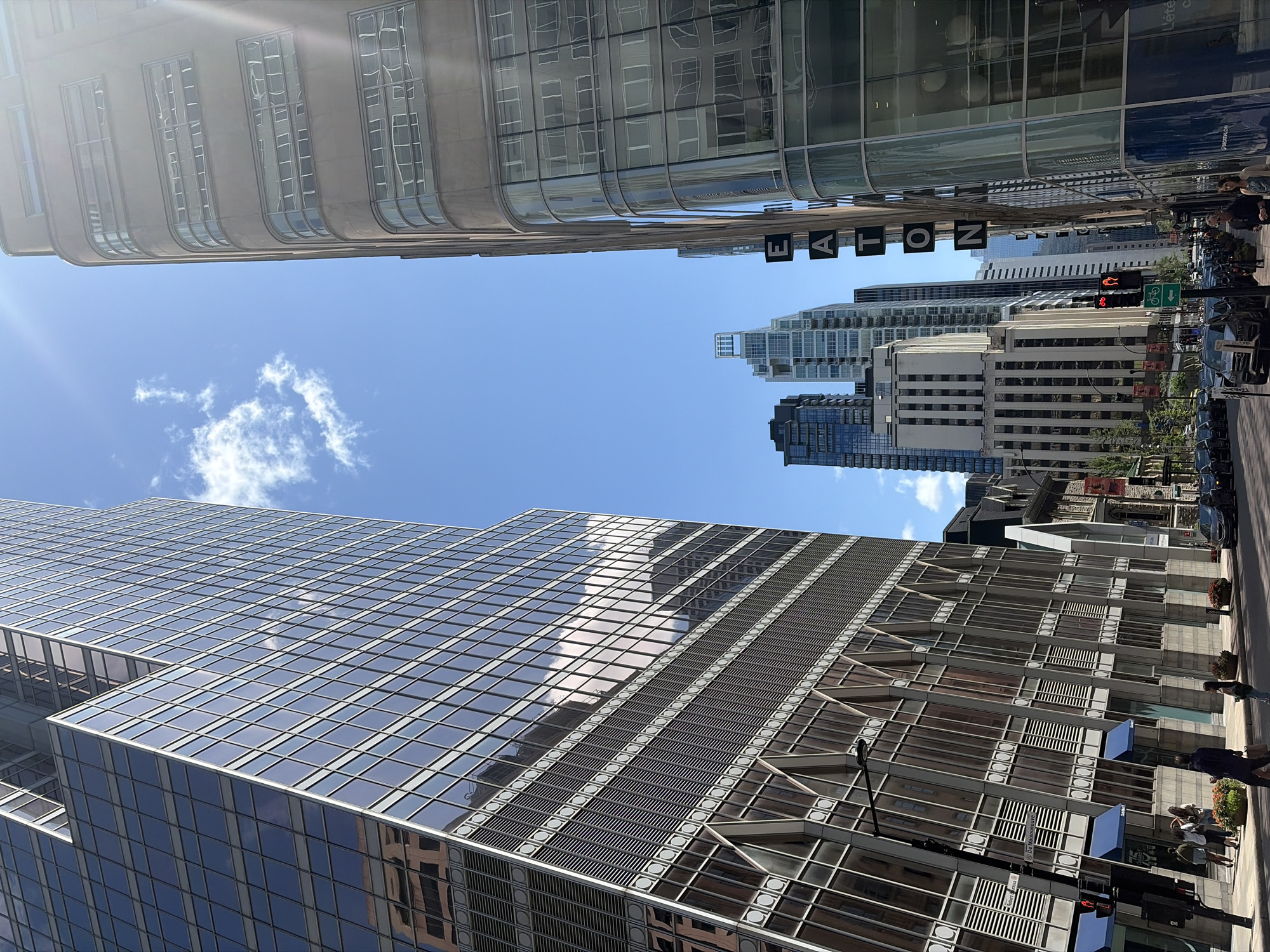 In an urban landscape under a bright, sunny sky with scattered white clouds, a street scene unfolds. The perspective is upward, looking towards several tall buildings. On the left, a modern skyscraper with a glass facade dominates the view, its reflective surface mirroring the sky and other buildings. Its angled design creates a striking visual effect. In the center, a line of various buildings stretches into the distance, some with darker glass and others with lighter facades, including a notable building with vertical white stripes. On the right, another building with a curved, beige facade and horizontal window bands is visible, with the word "EATON" vertically spelled out on its side in black letters. The street below is lined with parked cars and a few pedestrians. Traffic lights and street signs are present, indicating a city intersection. The overall impression is a bustling urban environment on a clear day.
