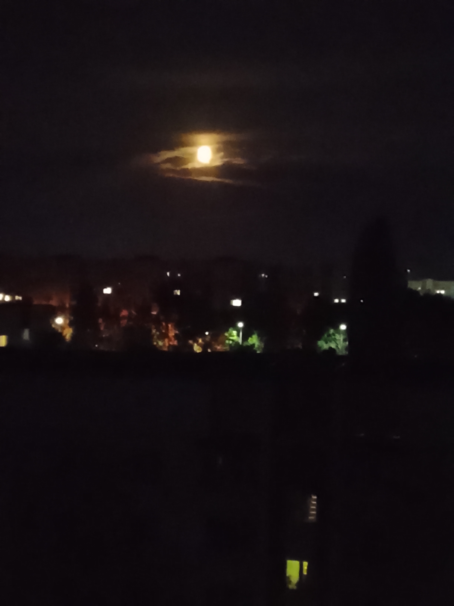 A nighttime photograph captures the full moon peeking through clouds over the city of Zhlobin, Belarus. The moon, a bright yellow orb, is partially obscured by wispy, dark clouds, creating a dramatic effect against the dark sky. Below, the cityscape is dimly lit by scattered streetlights and illuminated windows of buildings and houses, creating a soft glow on the horizon. Silhouetted trees and buildings are visible in the foreground and midground, suggesting an urban or suburban environment. The overall impression is one of a calm, quiet night.