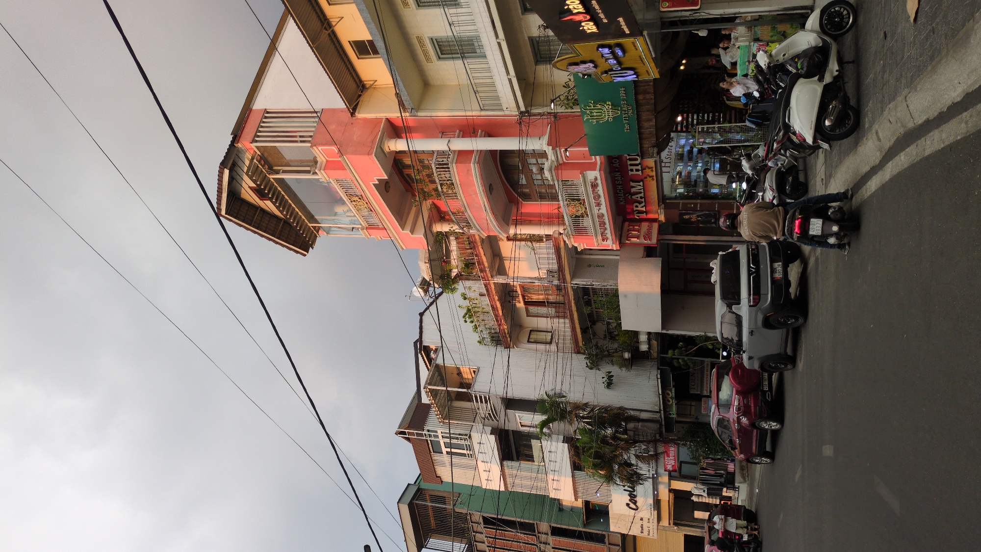 The image depicts a street scene in Da Lat, Vietnam. The street is lined with buildings of varying heights and architectural styles, some featuring balconies adorned with plants. Power lines are strung across the sky, visible against a light-colored sky. The street is occupied by cars and motorbikes, some parked and others with riders. The storefronts have signs in Vietnamese, some partially visible. One sign reads "STUDIO HOTEL KHÁCH SẠN ÁO TH TRẦM HƯ." Another sign advertises "The VINTAGE 1994 DALAT." People are present on the sidewalks and within the shops. The lighting suggests it is either early morning or late afternoon.