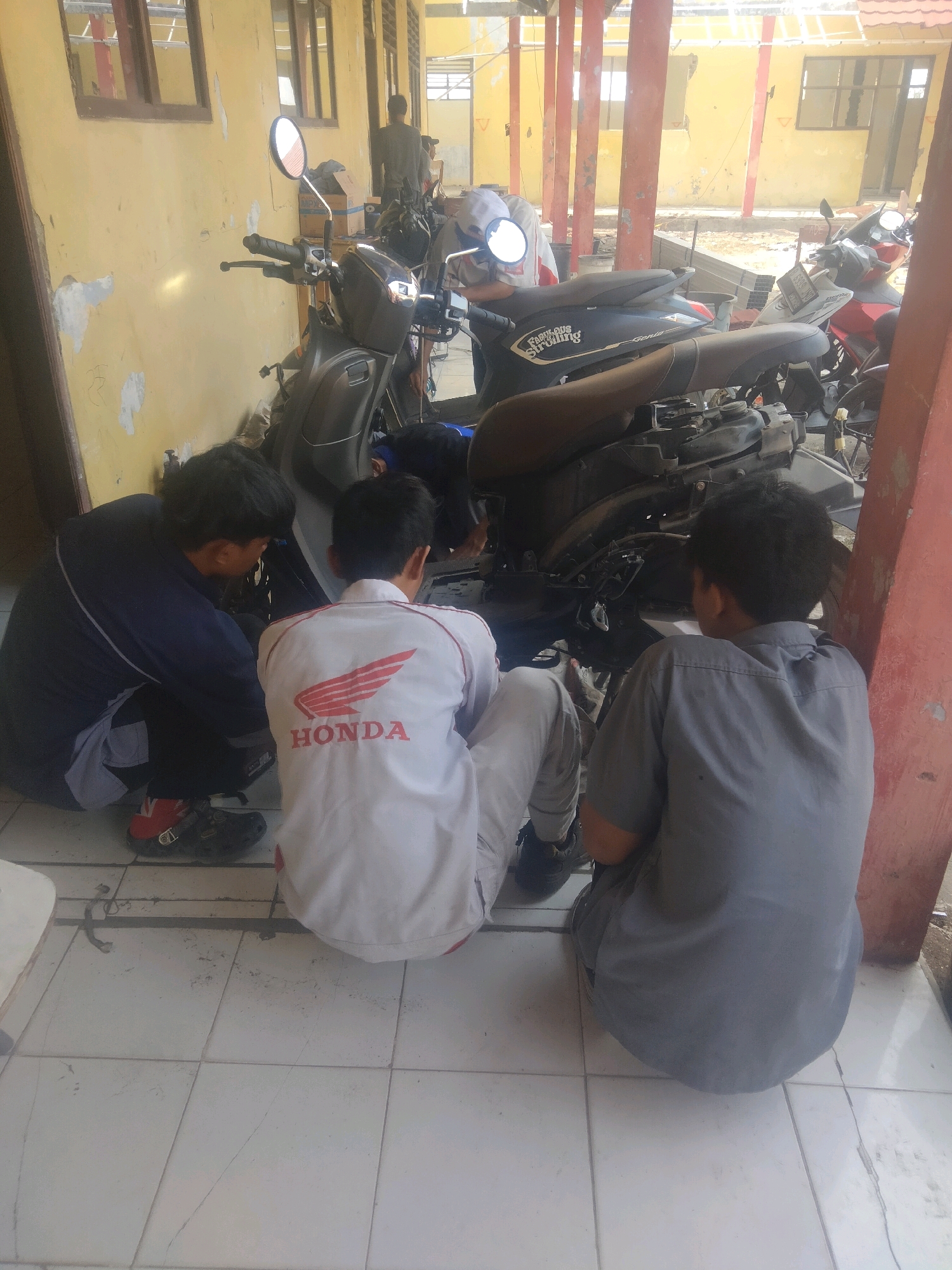 Three young males are crouched on a tiled floor next to several parked motorbikes. The setting appears to be an outdoor area with a yellow, weathered building in the background, supported by red pillars. One individual in the foreground, wearing a white jacket with a red Honda logo, is facing away from the camera. To his left, another individual in a dark blue jacket is also facing away. To the right, a third individual in a grey shirt is visible from the back. The motorbikes are in various states of assembly or disassembly, with exposed engines and parts. The overall atmosphere suggests a workshop or repair area. The image was taken in Suka Sari, Indonesia.