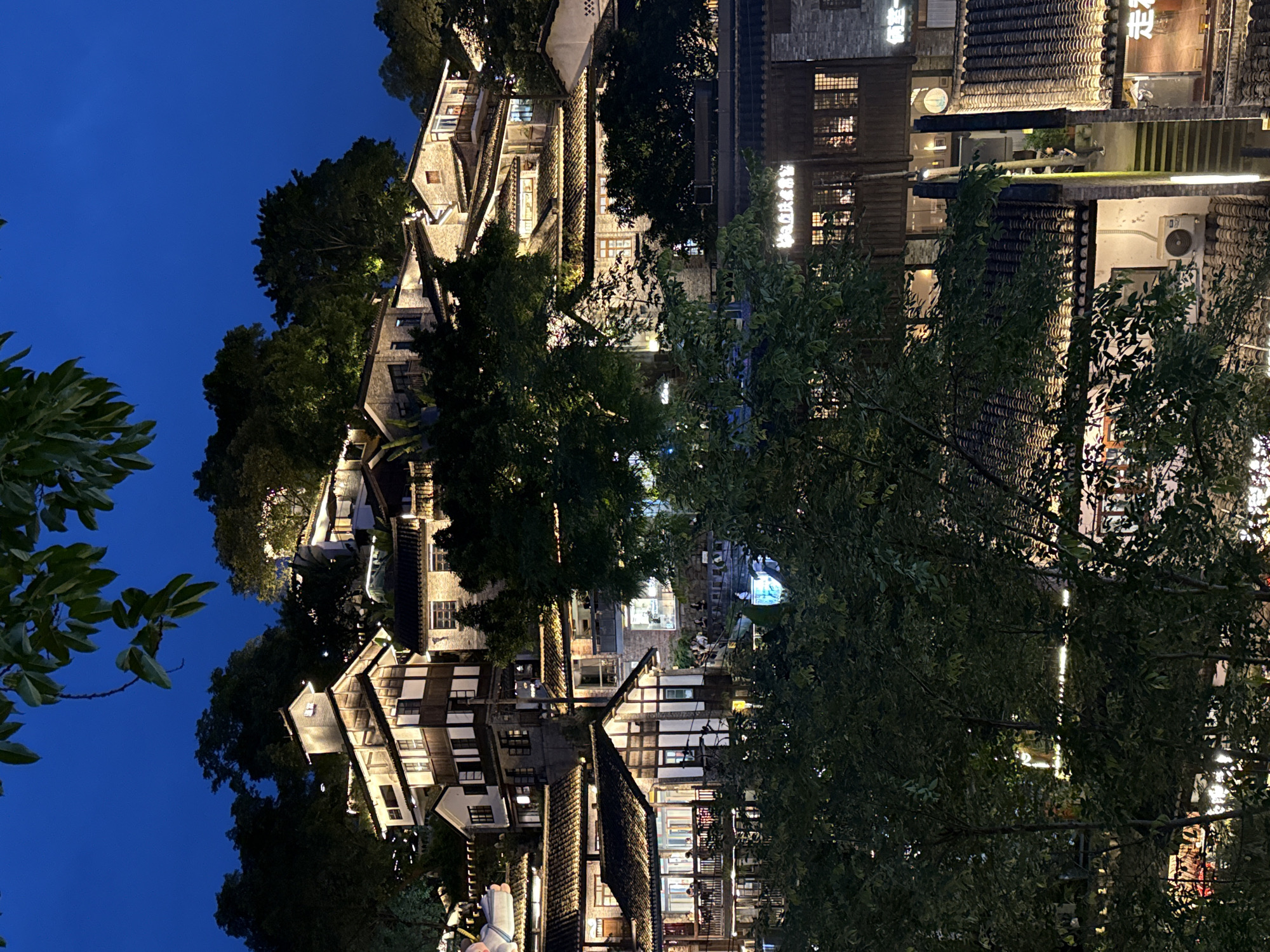 The image depicts a nighttime scene in Nan'an District, China. Buildings with illuminated windows are nestled amidst lush trees under a deep blue sky. The architecture appears traditional with dark tiled roofs. The overall atmosphere is calm and serene.