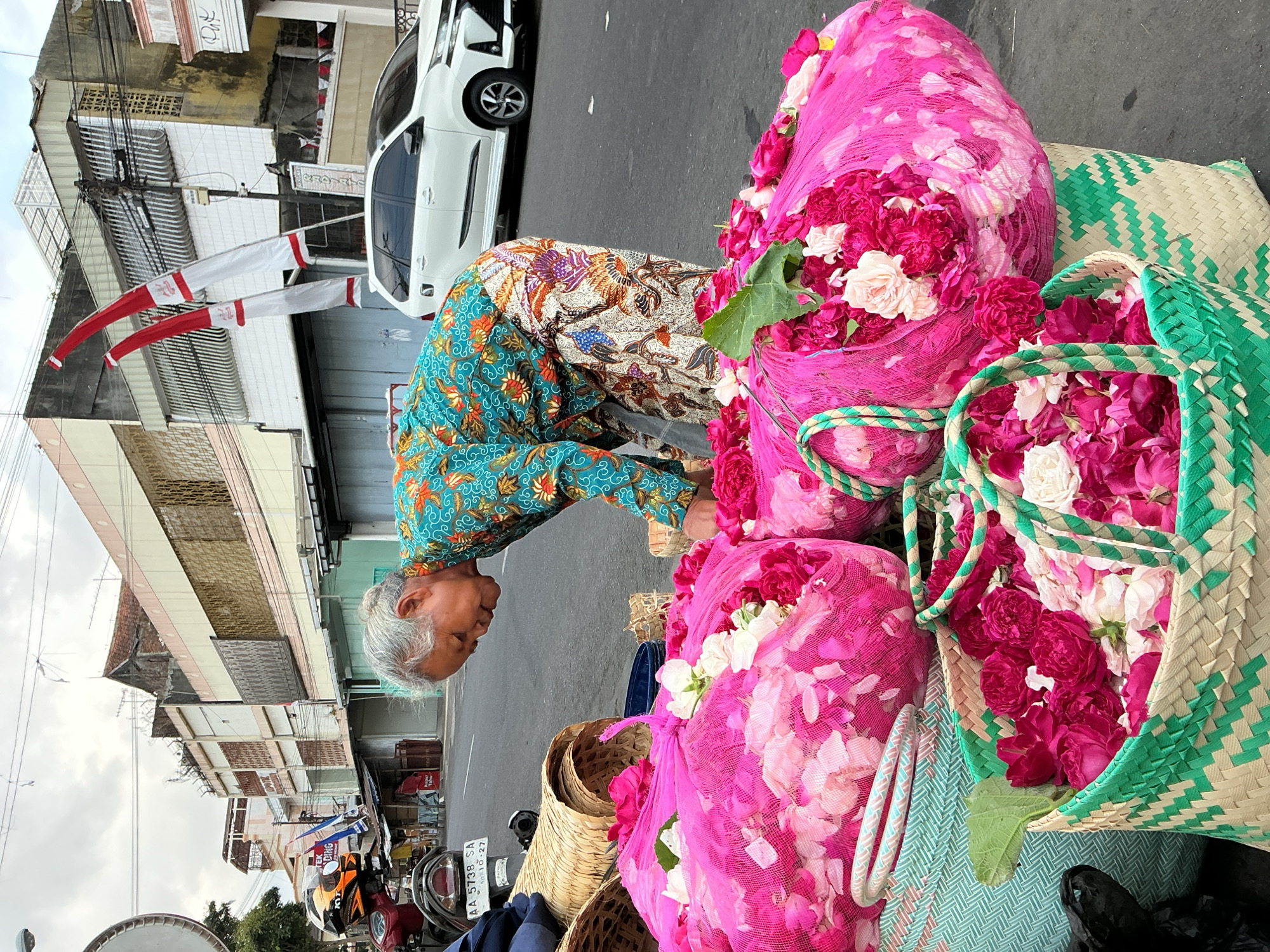 A street scene in Magelang, Indonesia, features a woman with gray hair wearing a colorful batik-patterned shirt and pants, bending over to arrange a large amount of pink and white flowers. The flowers are contained in two large, pink mesh bags and a woven basket. Behind her, a white SUV is parked on the asphalt road. The background shows a row of buildings with white and brown facades, and two red and white flags are visible on poles. A motorcycle is parked to the left of the frame. The ground is asphalt, and there are other woven baskets in the foreground. The sky is overcast.