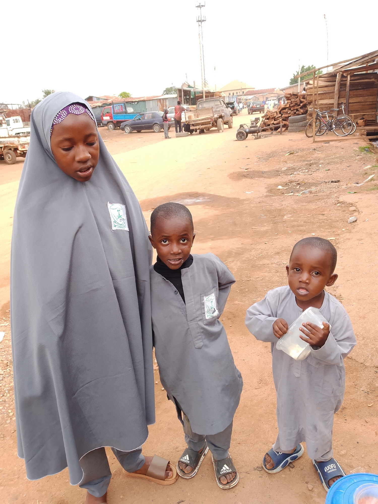 The image shows three children and a young woman standing in an outdoor marketplace. The young woman on the left wears a grey hijab and a long grey garment with a green emblem. She appears to be speaking. A young boy in the center, wearing a grey tunic over a black shirt, looks directly at the camera with a slight smile. To the right, another young boy in a similar grey tunic holds a clear plastic cup filled with a light-colored substance. He also looks at the camera. Both boys are wearing sandals.
In the background, the marketplace is visible. There are several vehicles, including trucks and cars. Piles of lumber and tires are stacked, suggesting it might be a construction or wood market. Buildings with corrugated metal roofs line the background. The ground is dirt, characteristic of a rural or less developed area. The sky is overcast, indicating a cloudy day. The overall impression is that of a casual, everyday scene in a community marketplace. There is no discernible text in the image, except for the emblems on the young woman's garment and logos on the children's sandals.