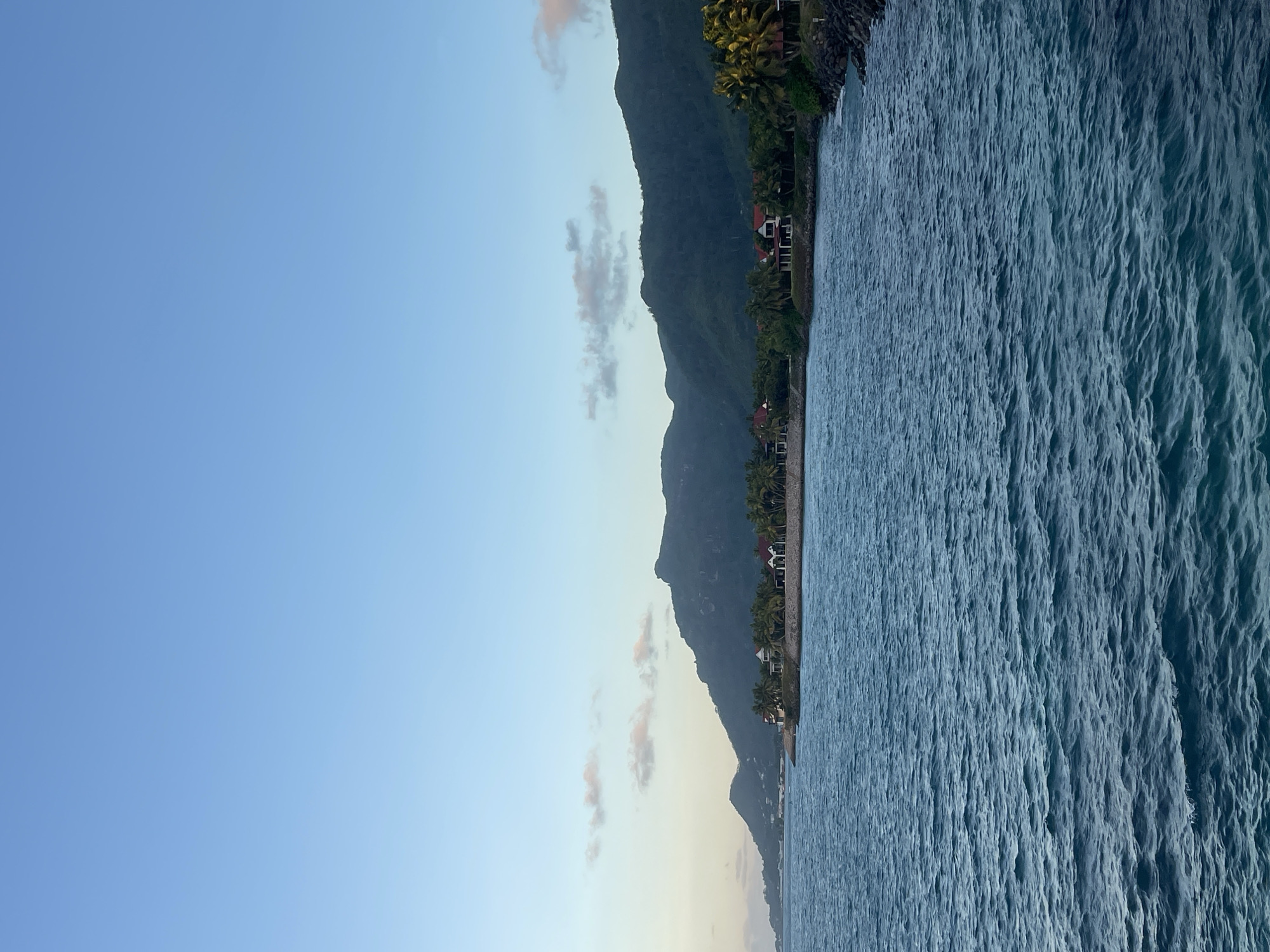 The image shows a coastal view of Victoria, Seychelles, at dusk. The foreground is dominated by the deep blue, rippling ocean. Along the coastline, lush green vegetation, including palm trees, can be seen, interspersed with a few residential buildings featuring red-tiled roofs. In the background, a large, tree-covered mountain range rises, its slopes darkening as the light fades. The sky above is a clear, pale blue, with a few wispy, pink-tinged clouds near the horizon. The scene depicts a tranquil coastal environment with a blend of natural landscape and human habitation.