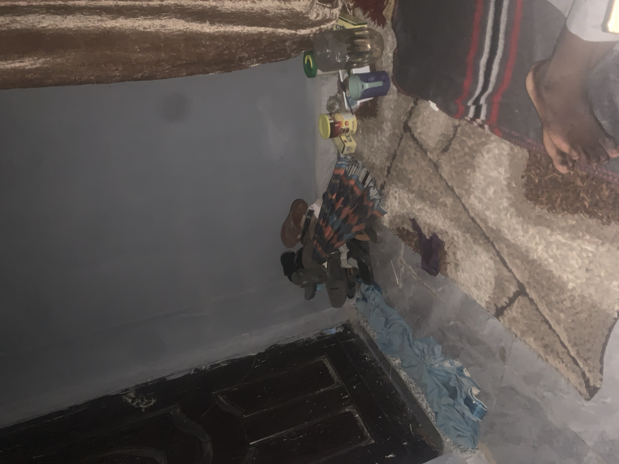 The image depicts an indoor scene, possibly a room in Jalingo, Nigeria. A dark wooden door with panels is visible on the left side of the frame. In front of the door, a pile of light blue cloth is lying on the floor. Adjacent to the cloth, on the right, is a collection of shoes, including several pairs of sandals and one pair of black shoes.
Further to the right, a multi-colored umbrella is placed on the floor, partially folded. Scattered around the umbrella are several objects: a yellow container with red lettering, a small white box, a blue cup with a handle, and a large clear glass jar filled with a liquid and a green lid.
In the background, a beige curtain hangs on the right side of the image. The walls are painted a dark gray. The floor appears to be tiled and covered in some areas with a textured rug. In the lower right corner of the image, a person's bare feet and legs are visible, resting on the rug. The person is covered by a dark blanket with horizontal red, white, and black stripes. The overall impression is of a cluttered domestic space.