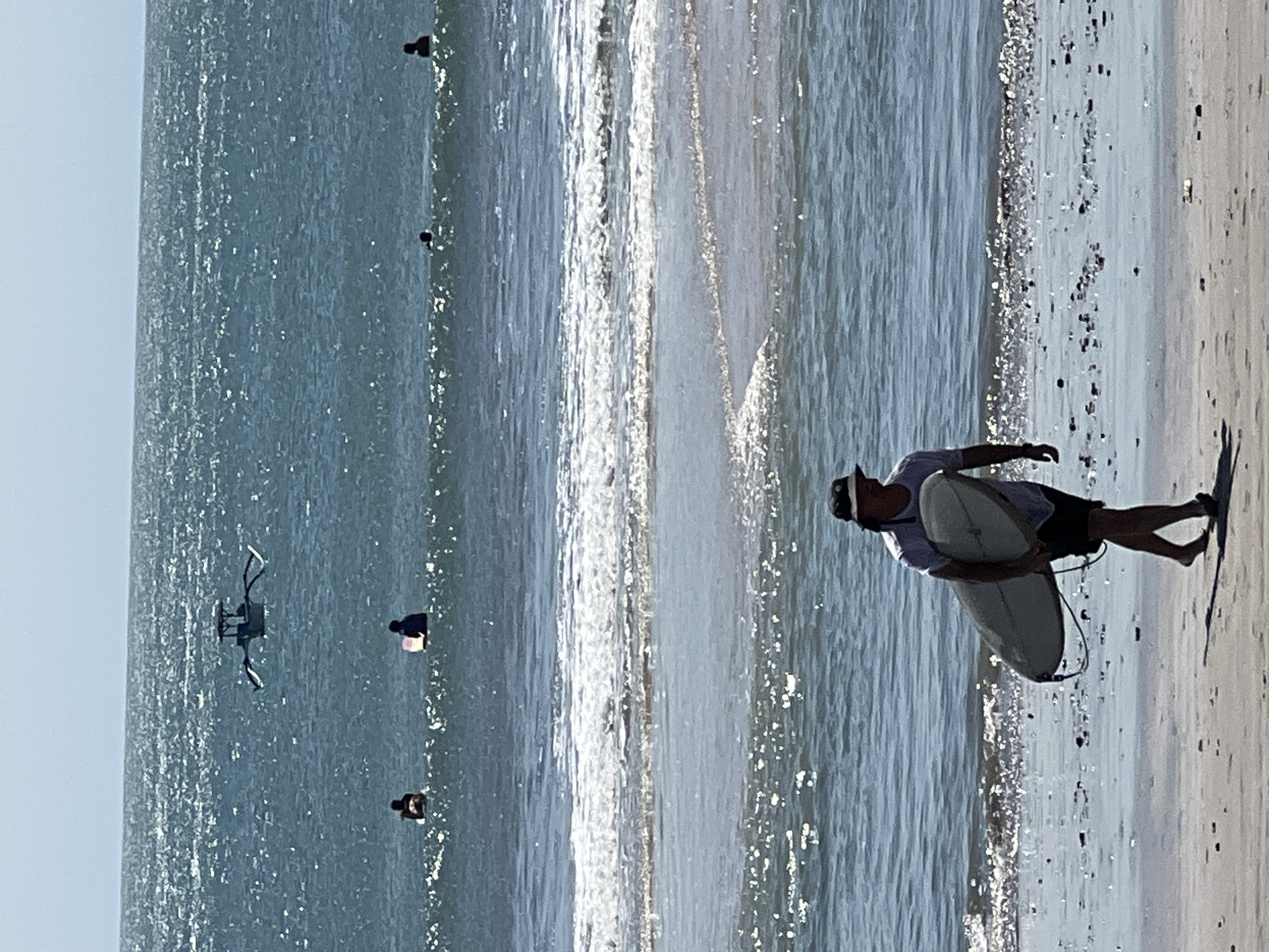 The image depicts a sunny day at Kuta Beach, Indonesia. A man wearing a hat, t-shirt, and shorts walks along the sandy shore, carrying a surfboard under his arm. In the ocean, several people are visible in the water, some appearing to be surfers or swimmers. A small boat with outriggers is also present in the distance. The water is blue with visible waves and a shimmering surface due to the sunlight. The beach is composed of sand with some scattered debris.