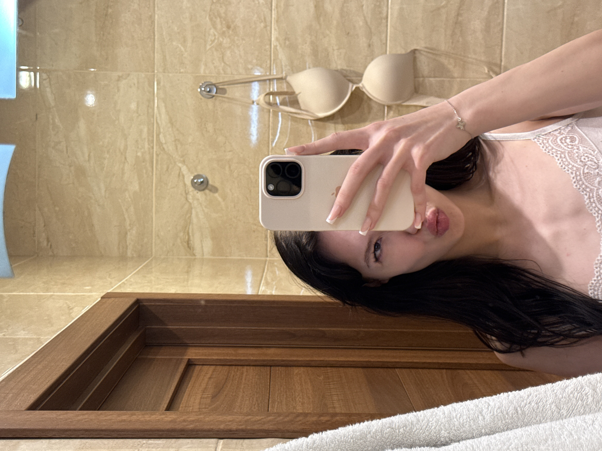 In a bathroom with beige tiled walls and a wooden door, a young woman takes a selfie in a mirror. She is holding a light-colored smartphone to her face, obscuring half of it, and making a pouty expression. She has long, dark hair and is wearing a white lace camisole. A light-colored bra is hanging on hooks behind her. A white towel is visible on the left side of the image. The lighting suggests an indoor environment.