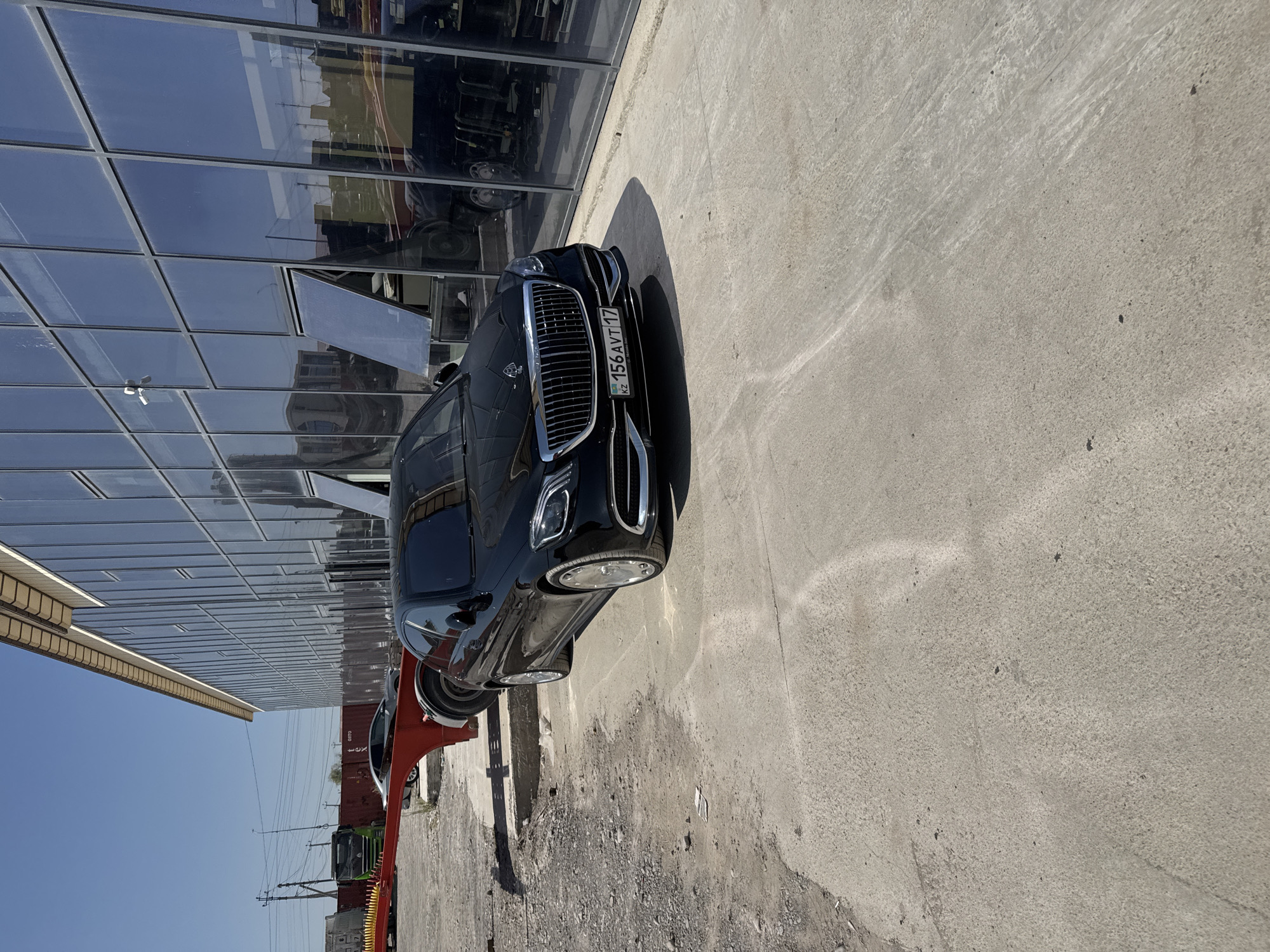 A black Mercedes-Maybach sedan is parked on a concrete surface in front of a modern building with a glass facade in Shymkent, Kazakhstan. The building appears to be an industrial or commercial establishment, with large windows reflecting the sky and surrounding objects. To the left of the car, a red trailer frame is visible, suggesting some form of transportation or construction equipment is present in the area. The ground is a mix of concrete and gravel. The sky is clear and blue, indicating a sunny day. The license plate of the car reads "KZ 156 AVT 17". There are no people visible in the image. The scene is a straightforward depiction of a vehicle parked in an urban or peri-urban environment.