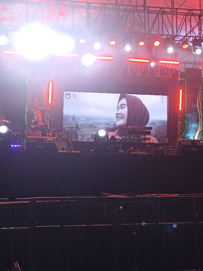 This media file depicts a lively outdoor stage event taking place at night in Kecamatan Pujut, Indonesia. The central feature is a large LED screen displaying a close-up image of a young woman smiling joyfully, wearing a dark head covering (hijab). The background behind her on the screen appears to be an open, possibly natural or rural, landscape.
The stage is structured with an elaborate metal truss system overhead, from which numerous bright stage lights, in white and red hues, illuminate the area. These lights create a dynamic atmosphere, with some causing noticeable lens flare in the image. To the left of the main screen, a person, possibly a performer or crew member, is partially visible, obscured by bright lights, and appears to be wearing distinct attire.
Musical instruments are arranged on the stage, including a drum kit, a keyboard on a stand, and a microphone stand. Faintly visible text on the bass drum seems to read "Go Drum". On the top left of the LED screen, a logo featuring a stylized "Go" is present, and some partially obscured text, possibly "PONTIA", can be seen near the bottom of the screen.
The foreground is dark, revealing the top of a barrier or railing, hinting at an audience beyond. The overall setting, with its bright lights, musical equipment, and large display screen, suggests a concert, festival, or similar public performance or event. The time of day is clearly nighttime, enhancing the dramatic effect of the stage lighting.