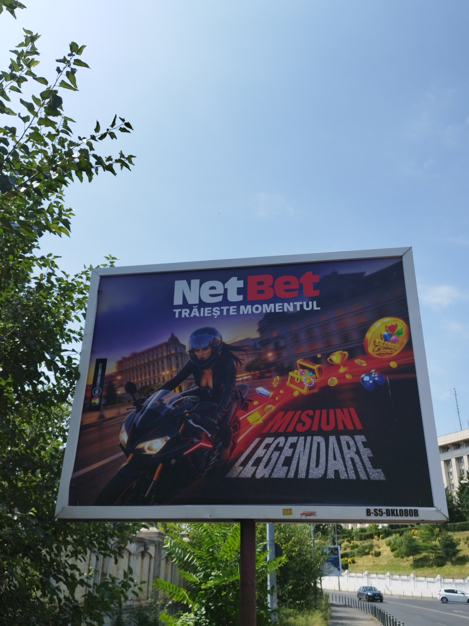 The image shows a billboard advertising NetBet, a company that appears to offer online gambling or betting services. The billboard features a woman on a motorcycle in the foreground, with a cityscape in the background. Various game-related imagery, such as treasure chests, coins, and trophies, are also depicted. Text on the billboard reads "NetBet Trăiește Momentul Misiuni Legendare," which translates to "NetBet Live the Moment Legendary Missions." The billboard is situated along a road in Bucharest, Romania, as indicated by the presence of cars on the road and typical Romanian architecture in the background. The sky is clear and blue, with some foliage from nearby trees partially obscuring the billboard on the left side of the image. The billboard is mounted on a pole and is a prominent advertisement in an urban environment.