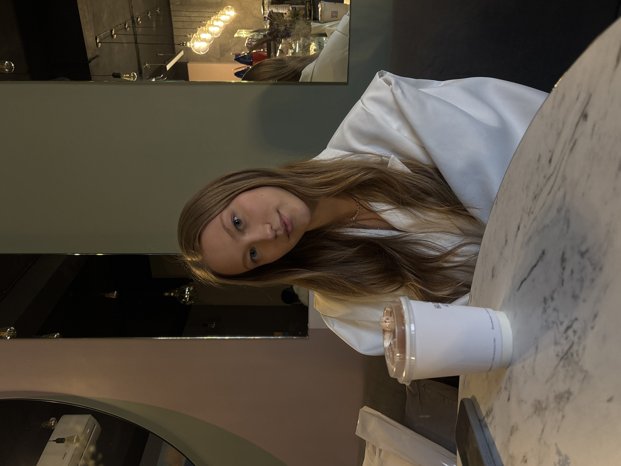 A young woman with long brown hair and blue eyes, wearing a white shirt, sits at a marble-topped table. On the table, there is a cup with a clear lid. Behind her is a mirror reflecting a bar with hanging light fixtures. The location is likely a cafe or bar in the city of Perm, Russia.
