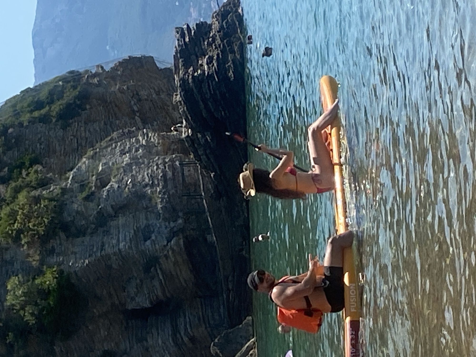 In a scene set against the dramatic, rocky coastline of Budva, Montenegro, two individuals are enjoying a sunny day on the water. A woman, wearing a bikini and a hat, is paddling a yellow stand-up paddleboard. She is lying on her stomach, with one leg bent and resting on the board, and holding a paddle. To her right, a man is also on a paddleboard, wearing sunglasses, a cap, and an orange life vest over black shorts. He is sitting on his board, facing the camera, with his hands resting on the board. Both individuals appear relaxed and are enjoying the clear, blue water that reflects the bright sunlight. The water is calm, with gentle ripples. In the background, the rugged cliffs are covered in sparse vegetation, suggesting a Mediterranean landscape. The overall mood is one of leisure and outdoor recreation on a pleasant day. The visible text on the paddleboard reads "FUSION."
