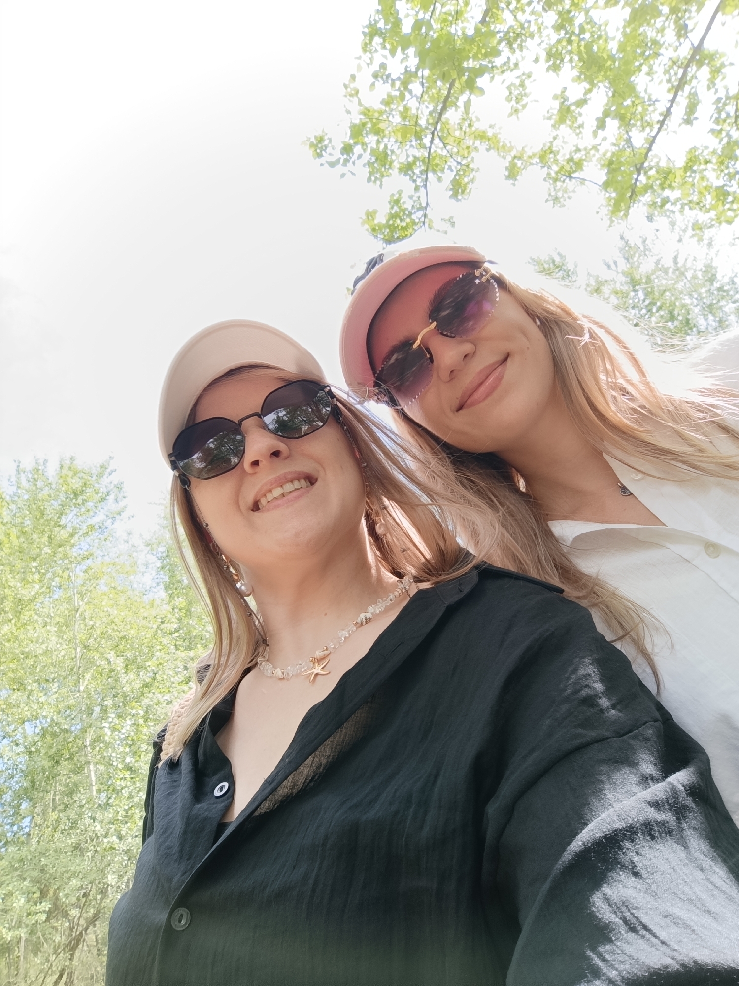 Two young women are captured in a low-angle, close-up selfie taken outdoors, possibly on a sunny day given the bright sky and dappled sunlight filtering through tree leaves overhead. The woman on the left wears a light-colored baseball cap and dark sunglasses. She has blonde hair and is smiling broadly, her teeth visible. She is wearing a black, collared shirt with visible buttons and a necklace featuring clear beads and a starfish charm.
The woman on the right is also wearing sunglasses, which appear to have a pinkish tint and a decorative gold accent on the sides. She has long, blonde hair and is wearing a pink baseball cap. Her expression is a gentle smile. She is wearing a white, collared shirt. The background consists of green tree foliage and a bright, blown-out sky, suggesting it is daytime. The overall mood appears cheerful and casual, as if the subjects are enjoying an outdoor activity together. No text is visible in the image.