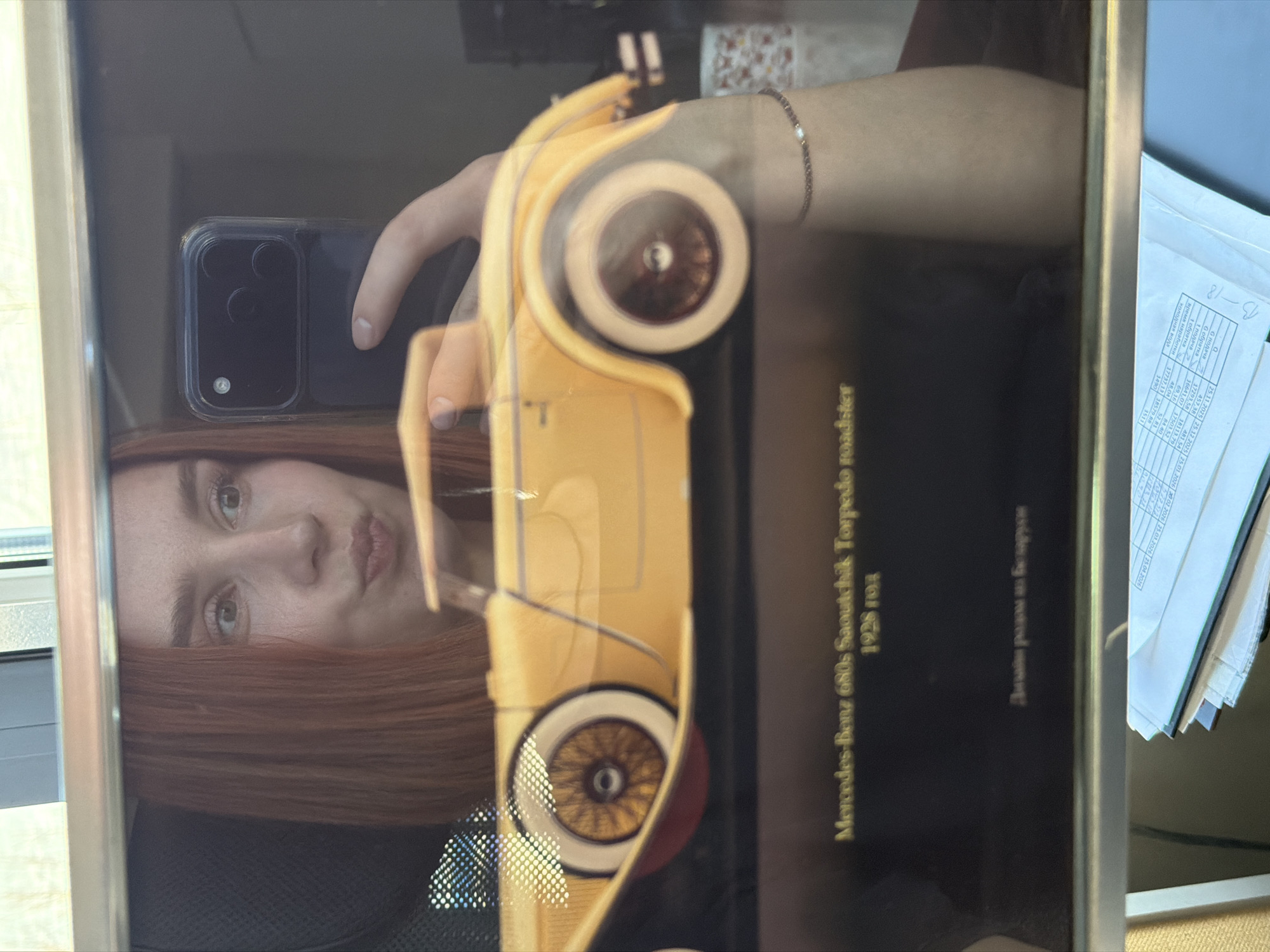 The image shows a woman taking a picture of a framed picture of a yellow Mercedes-Benz 680s Saoutchik Torpedo roadster from 1928. The text "Mercedes-Benz 680s Saoutchik Torpedo roadster 1928 год" is visible. A hand wearing a bracelet is also visible, holding a smartphone. A stack of papers sits to the right. The photograph is taken indoors. There's no evidence in the image to indicate the location is the city of Chaikovsky, Russia.