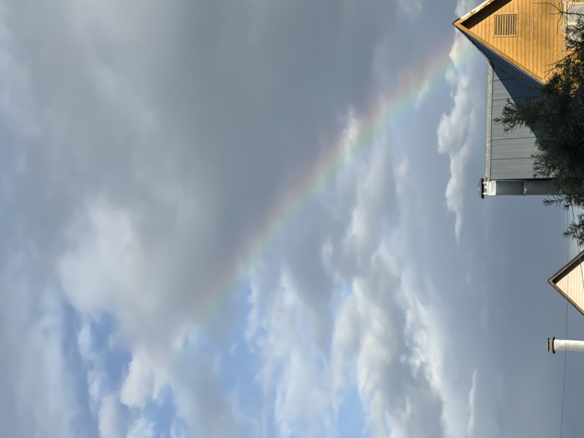 This is a vertical, outdoor, eye-level shot of a rainbow in Satino-Russkoe, Russia. A rainbow arcs across a partly cloudy sky, with the upper portion of the rainbow disappearing into dark gray clouds on the right side of the frame, and the lower portion extending into brighter, blue sky filled with white clouds on the left. The bottom of the image features the rooftops of two buildings. On the left, a silver-colored metal roof with a white chimney protrudes from the frame. To the right of this, a yellow wooden house with a peaked roof is visible. The rainbow is prominently featured in the sky above the buildings.