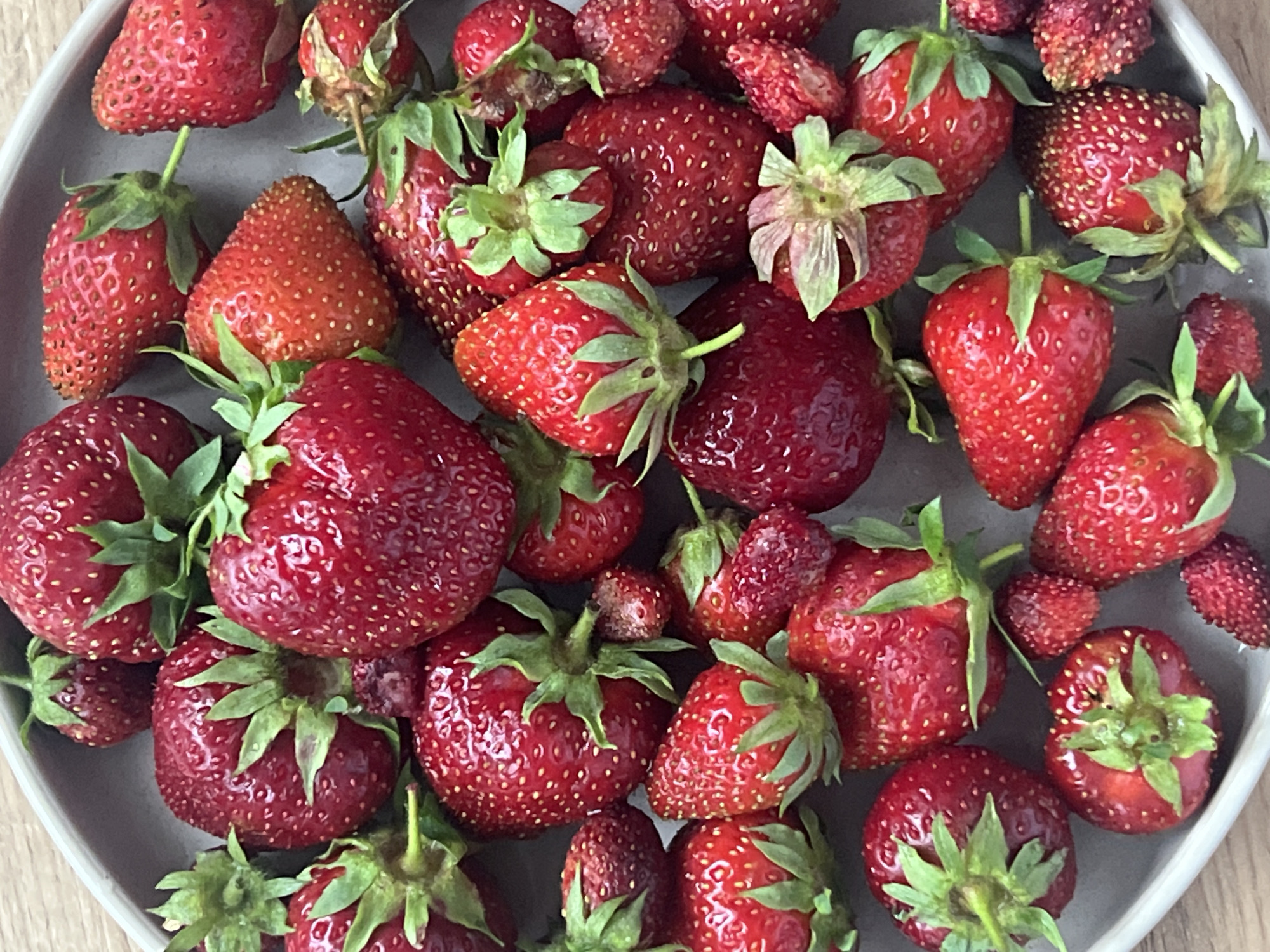 A close-up, top-down shot features a grey, shallow bowl filled with ripe strawberries. The strawberries are a vibrant red, with many showcasing their green stems and leafy calyxes. Some berries are larger and plumper, while others are smaller and more elongated. The surfaces of the strawberries are dotted with tiny seeds. The background is a light brown, textured surface, likely a wooden table. The lighting appears to be natural, casting subtle shadows on the fruit. The scene is devoid of people or any indication of location in Votkinsk, Russia, beyond the potential for the image to have been taken there.
