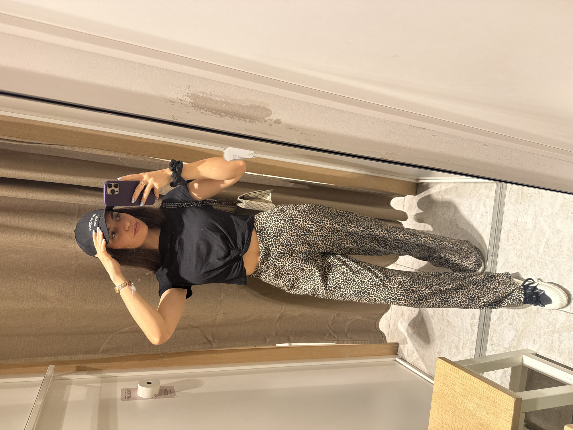 A young woman is taking a mirror selfie. She wears a black t-shirt knotted at the waist, leopard print pants, and a black baseball cap. She holds a purple phone. She is standing in a fitting room with a beige curtain and light-colored flooring. It is not possible to determine if the photo was taken in Plovdiv, Bulgaria from the given information.