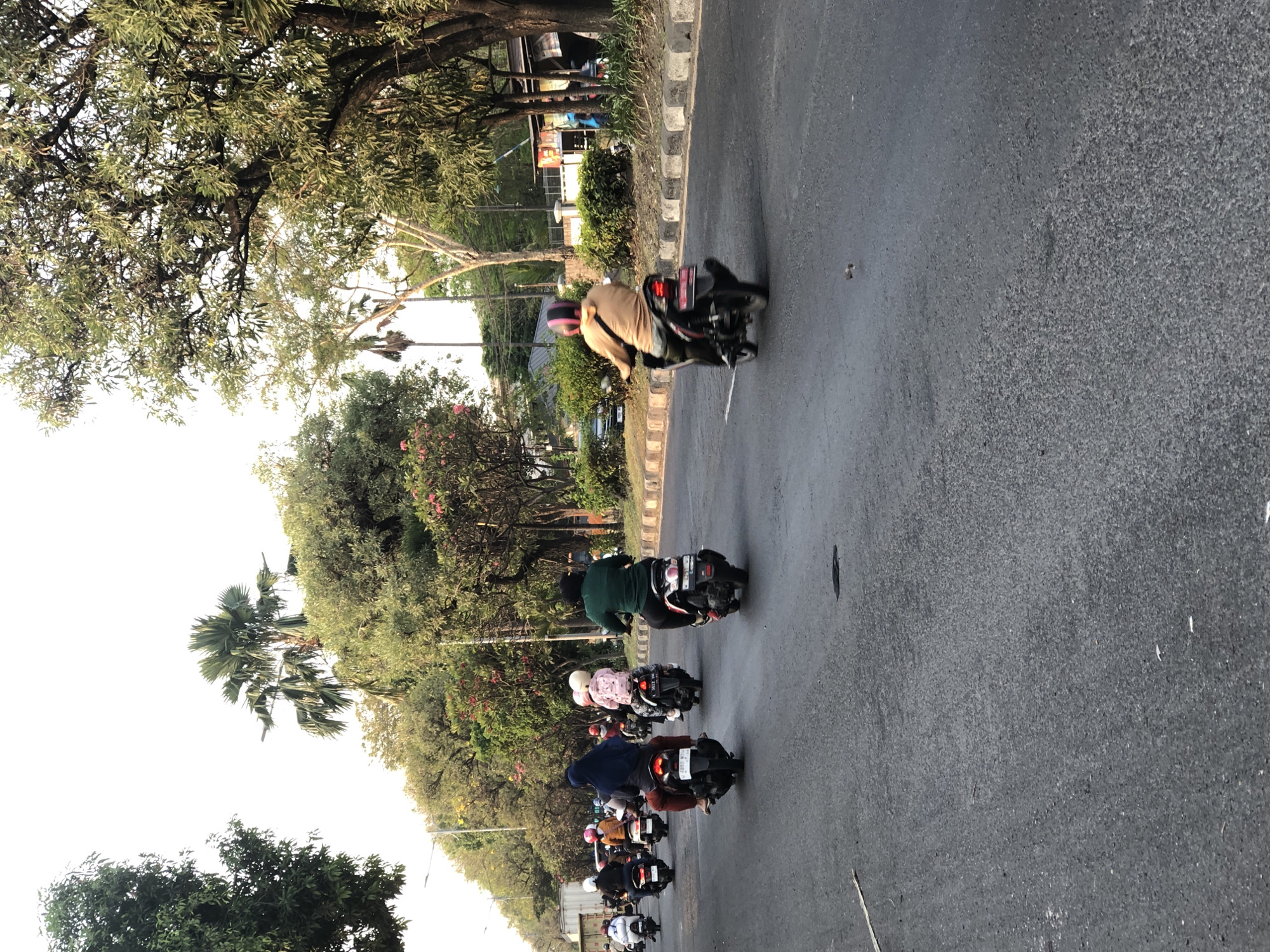 The image shows a street scene in Ngaban, Indonesia, with several people riding motorcycles. The road is paved and appears to be a two-lane road with a sidewalk lined with trees and some buildings in the background. The sky is overcast, suggesting it might be early morning or late afternoon. The motorcycles are of various makes and models, and the riders are dressed in casual clothing. Some riders are wearing helmets, while others are not. The overall impression is a busy street with a lot of motorcycle traffic.