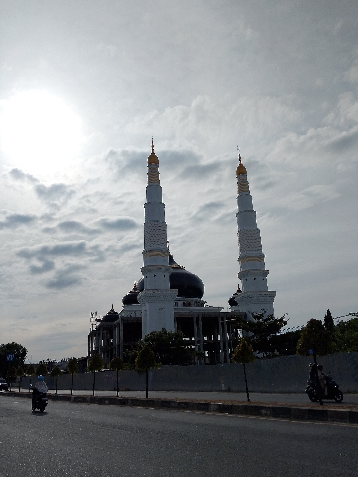 The image shows a mosque with two tall minarets, likely under construction, in Pantegajah, Indonesia. The sky is cloudy, with the sun partially obscured by clouds, creating a bright haze. In the foreground, a paved road runs horizontally across the frame, with a sidewalk and a low barrier separating it from a construction site. A lone motorcycle with a rider wearing a light-colored shirt and a blue helmet is visible on the road, traveling away from the viewer. Another motorcycle with two riders is further down the road, also moving away. Several young trees are planted along the sidewalk. The mosque itself has a large black dome and white walls, with intricate detailing on the minarets. Scaffolding is visible on parts of the mosque structure, indicating ongoing construction work. The overall scene depicts a partially completed religious building within an urban environment on a cloudy day.