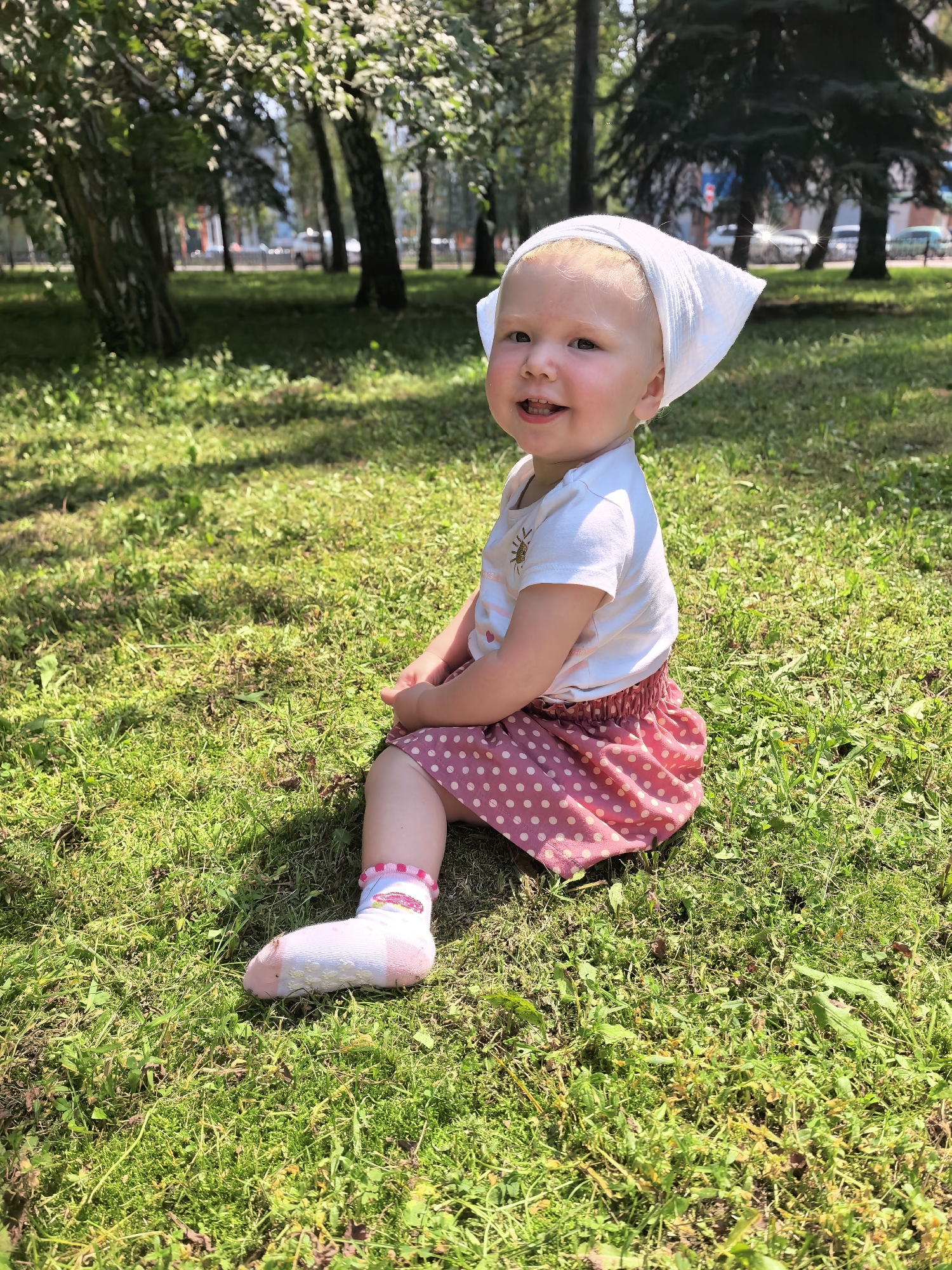 An infant with blonde hair, wearing a white bandana, a white t-shirt with a sun graphic, a pink polka dot skirt, and white socks with pink trim, sits on a grassy lawn in a park. The child is looking towards the camera with a smile and appears to be around one to two years old. The background shows trees, a street with cars, and buildings, suggesting an urban park setting. The lighting indicates a sunny day.
