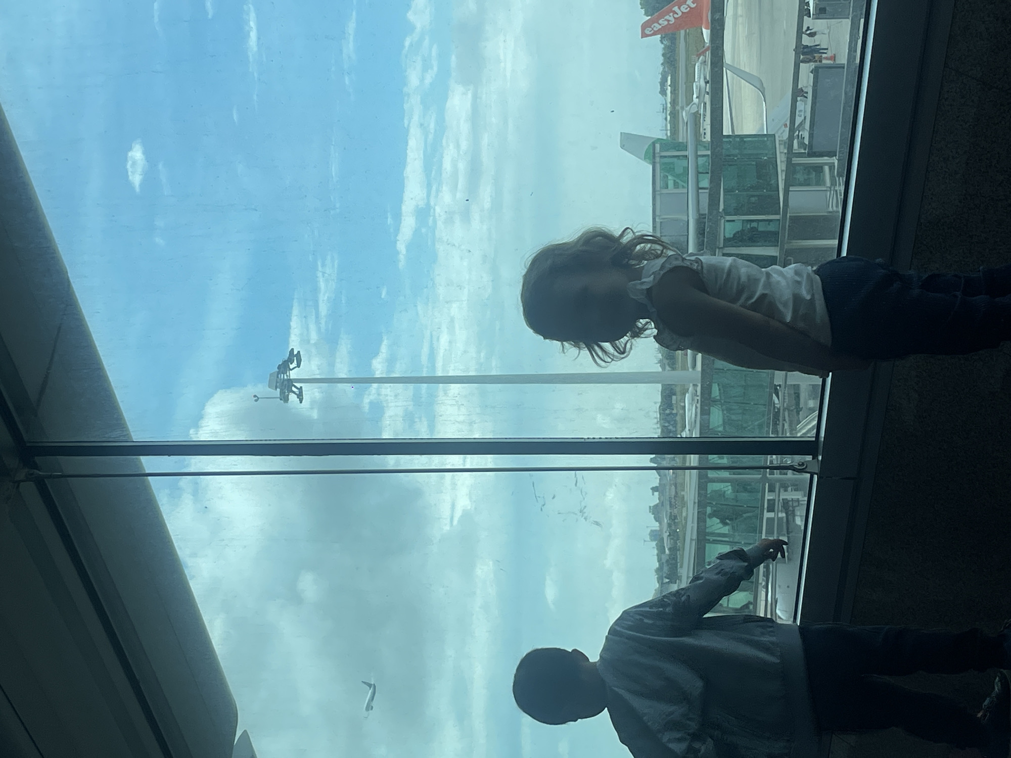 The image, correctly oriented, shows two young children standing by a large window, looking out onto an airport tarmac. A young girl, with curly hair and wearing a light top and dark trousers, is positioned on the right side of the frame, gazing intently outwards. To her left, a young boy, wearing a patterned top and dark trousers, is also looking out and has his right arm extended, pointing towards the scene outside.
The children are primarily silhouetted against the bright light from outside, making their facial features indistinct but highlighting their posture of observation. Outside the window, under a bright, cloudy sky, an airport apron is visible. An easyJet airplane, identifiable by its orange tail fin and "easyJet" logo, is parked on the tarmac. Various airport structures, possibly jet bridges or other terminal buildings, are also visible. The scene captures a moment of interest and wonder for the children as they observe the activity at what is identified as Moreira, Portugal, during the daytime with a bright, overcast sky.
