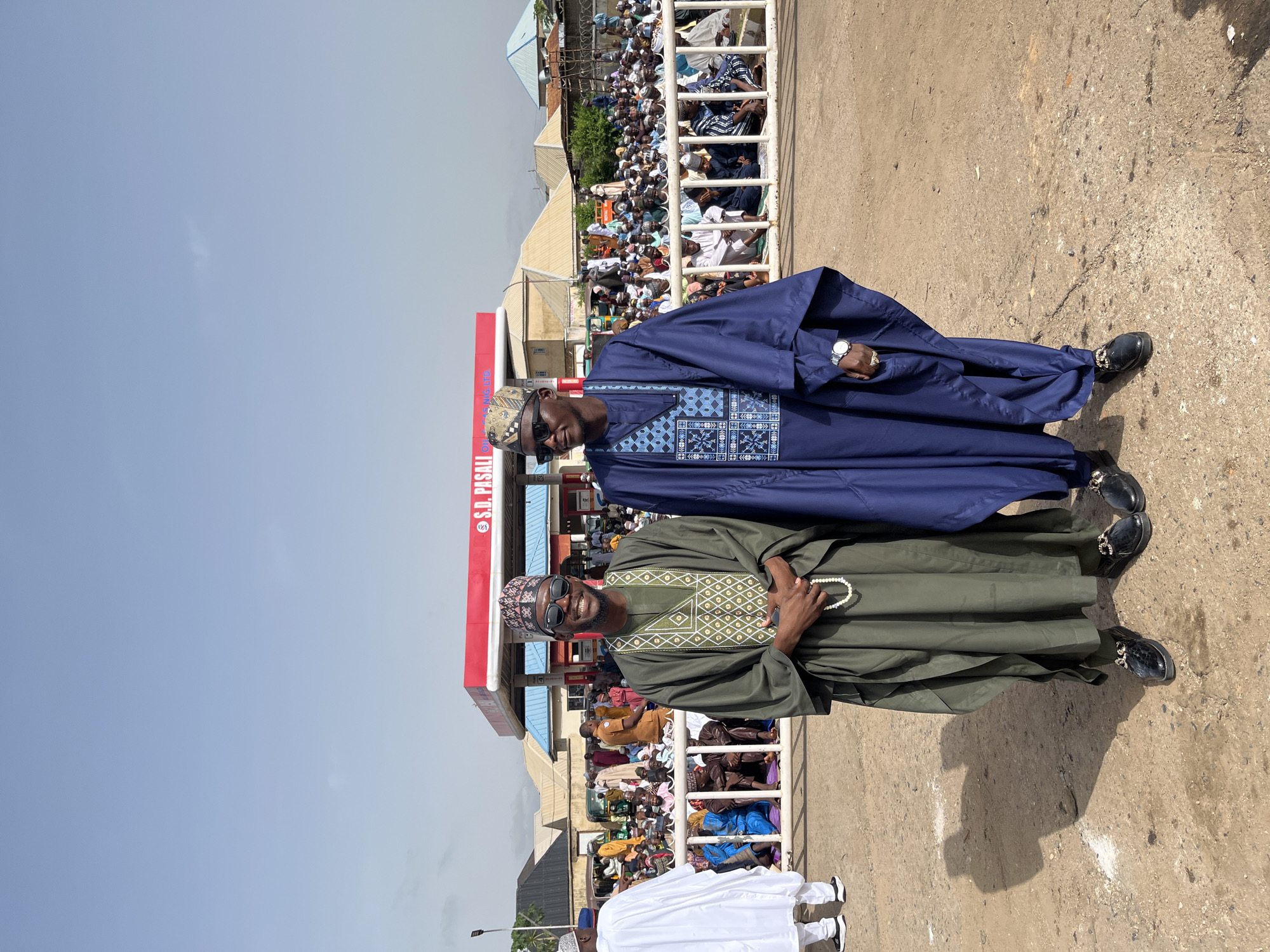 Two men are standing in front of an "S.D. Pasali" gas station, amidst a crowd of people beyond a white barrier. The man in the foreground is wearing an olive green agbada with patterned embroidery around the neck. He is holding a string of white prayer beads. The other man is wearing a dark blue agbada with similar embroidery. Both men are wearing matching caps, sunglasses, and black shoes with gold detailing. The sky is clear and pale blue. The location is presumed to be in Jos, Nigeria.