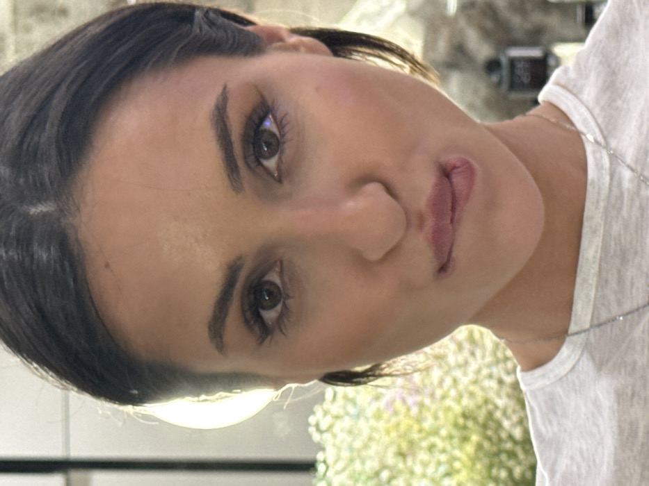 A woman faces the camera, tilted slightly. She has dark hair pulled back, accentuated eyebrows, and brown eyes with long lashes. Her skin appears smooth, and she wears a subtle lipstick. She wears a casual white T-shirt with a delicate silver necklace. Soft lighting creates a gentle glow on her face. In the background, there's a blur of greenery and glimpses of what appears to be a room setting.